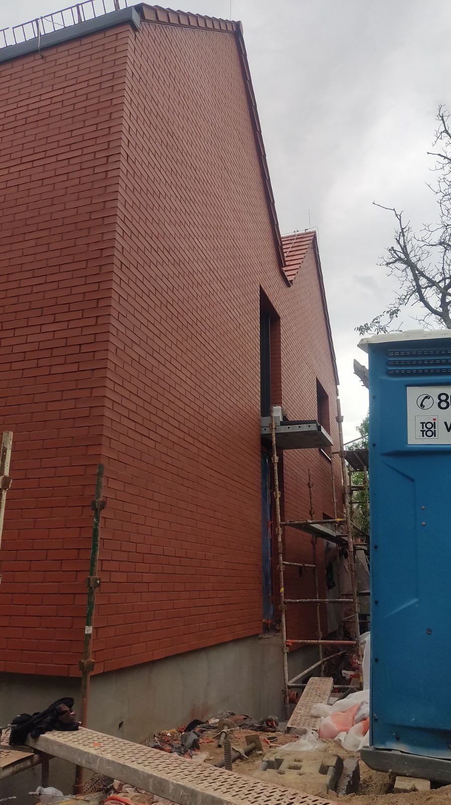 Nowo wybudowany budynek z cegły klinkierowej w odcieniach czerwieni, widoczne rusztowania i przenośna toaleta na placu budowy, pochmurne niebo w tle.