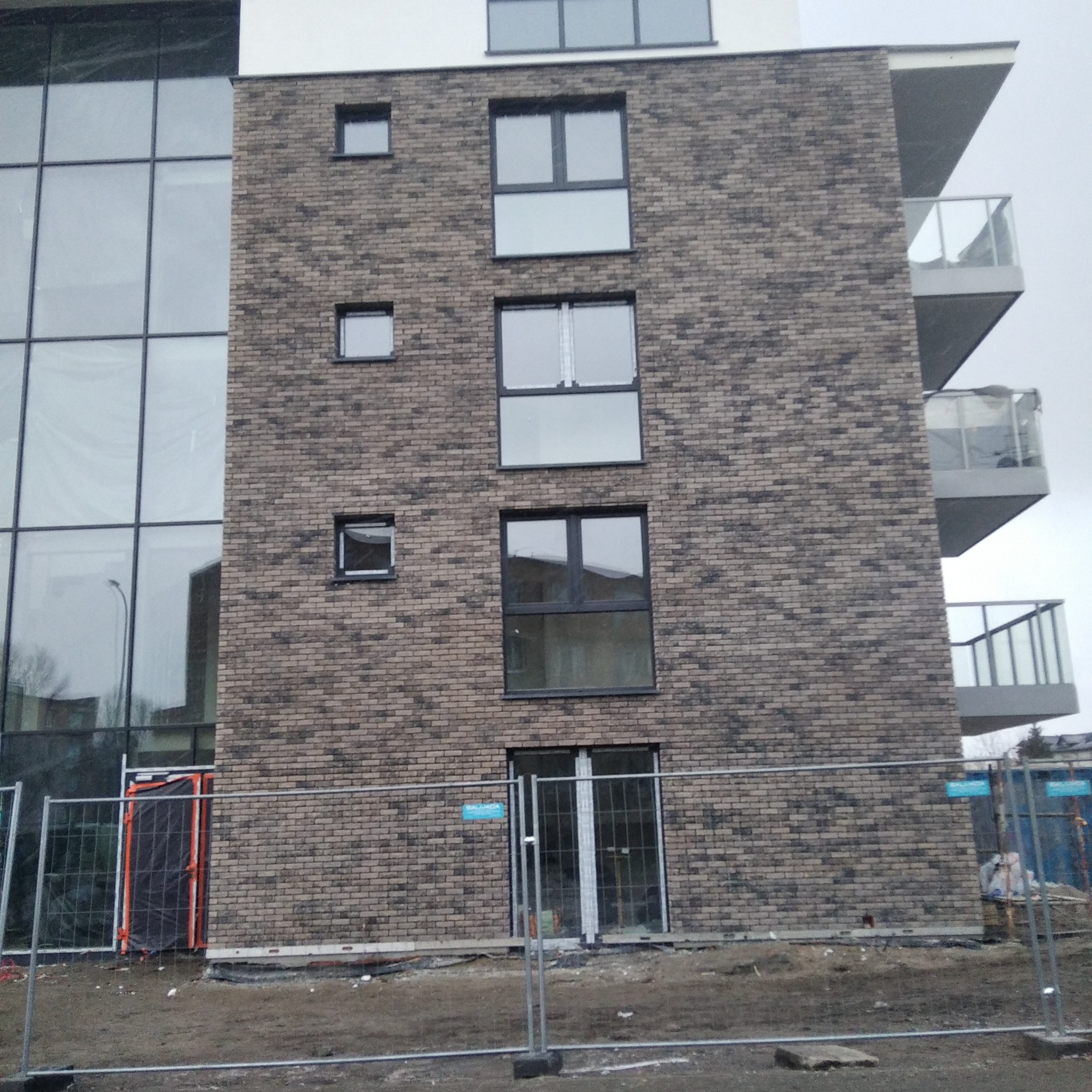 Elewacja budynku wielorodzinnego z cegły klinkierowej w trakcie budowy, widoczne okna, balkony z barierkami szklanymi oraz tymczasowe ogrodzenie budowy.
