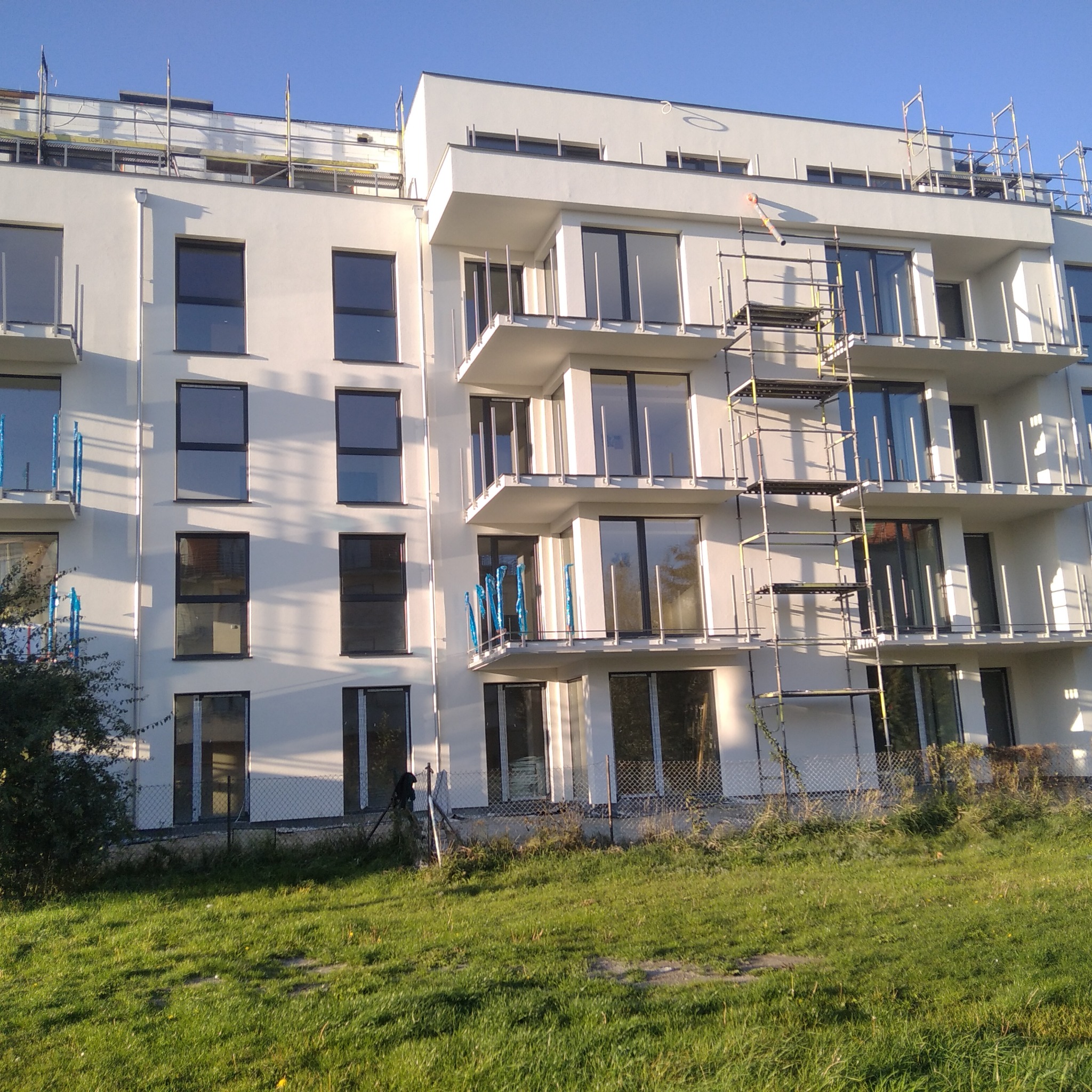 Nowoczesny, biały budynek wielorodzinny z balkonami, częściowo osłoniętymi folią, z rusztowaniami przy elewacji, otoczony zielonym trawnikiem.