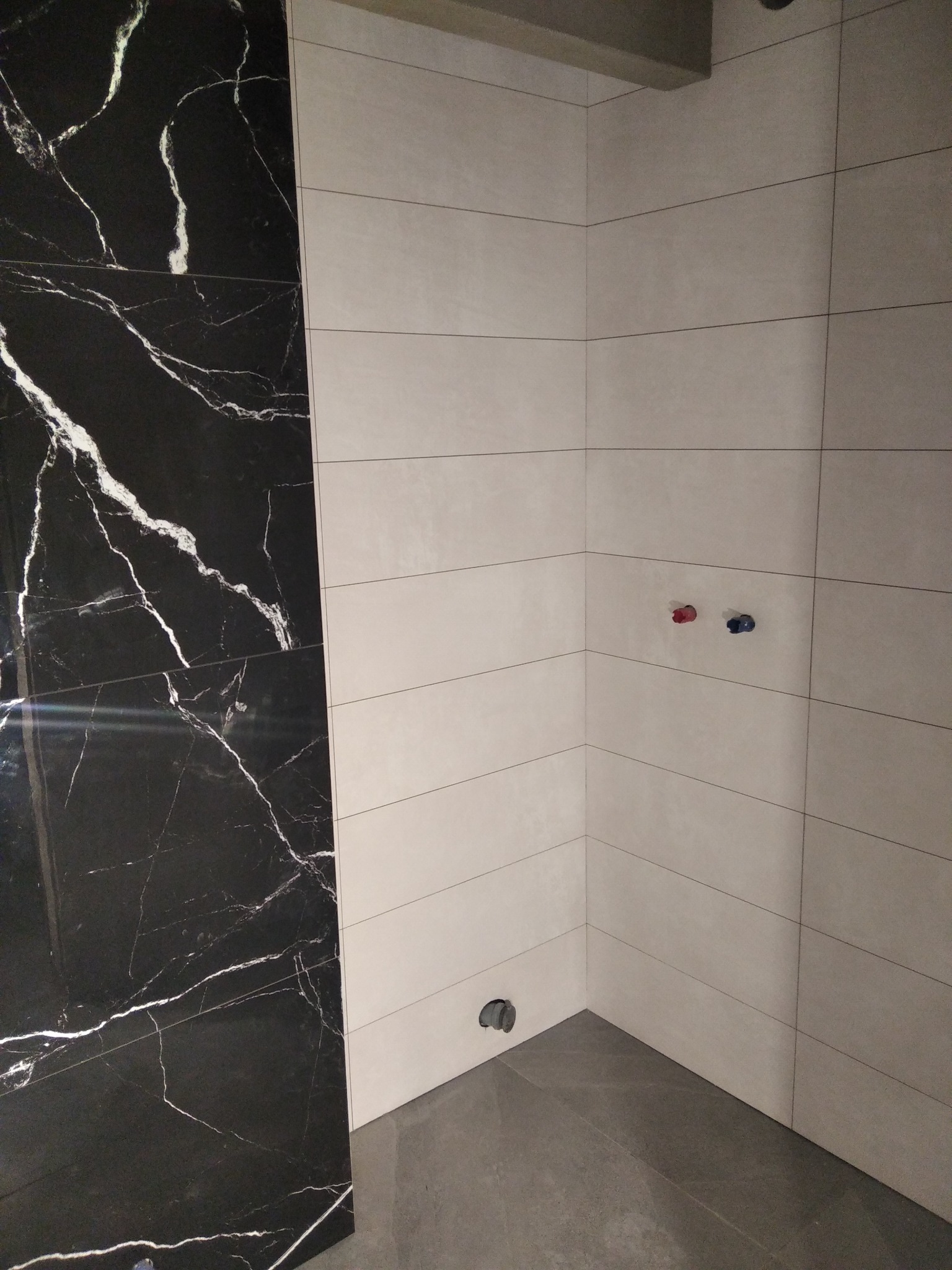 Fragment łazienki z wyłożonymi płytkami: ściana z czarnych płytek imitujących marmur i ściana z beżowych, poziomych płytek. Widoczne przyłącza wodne i odpływ pod prysznic.