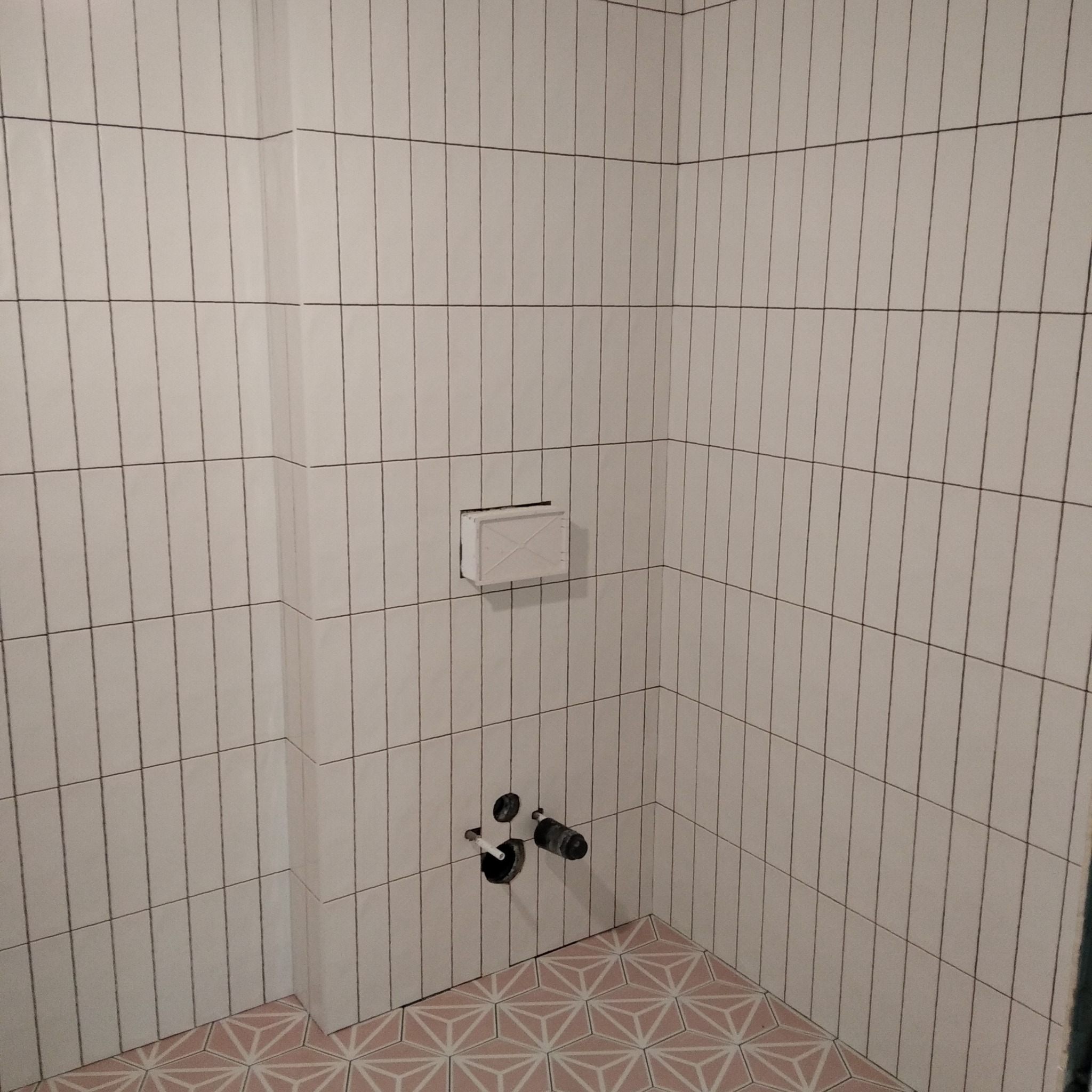 Wykończona łazienka z pionowo ułożonymi, wąskimi, białymi płytkami z ciemną fugą, dekoracyjna podłoga w geometryczne wzory w odcieniach różu i bieli, widoczne rury i otwór wentylacyjny.