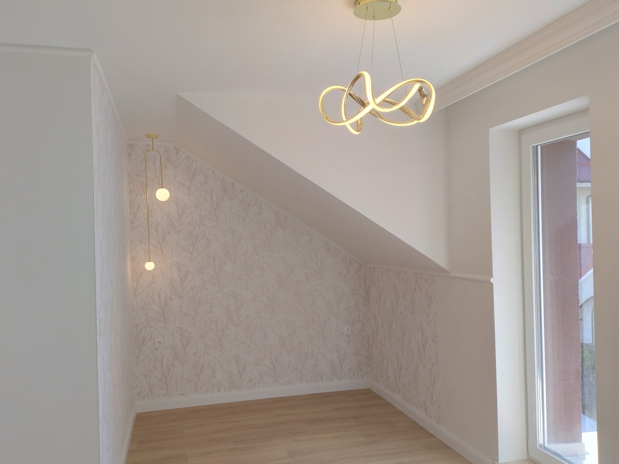 Pokój na poddaszu po remoncie: jasne panele, ściany w jasnym kolorze z delikatnym wzorem, nowoczesne lampy wiszące, okno z widokiem na zewnątrz.