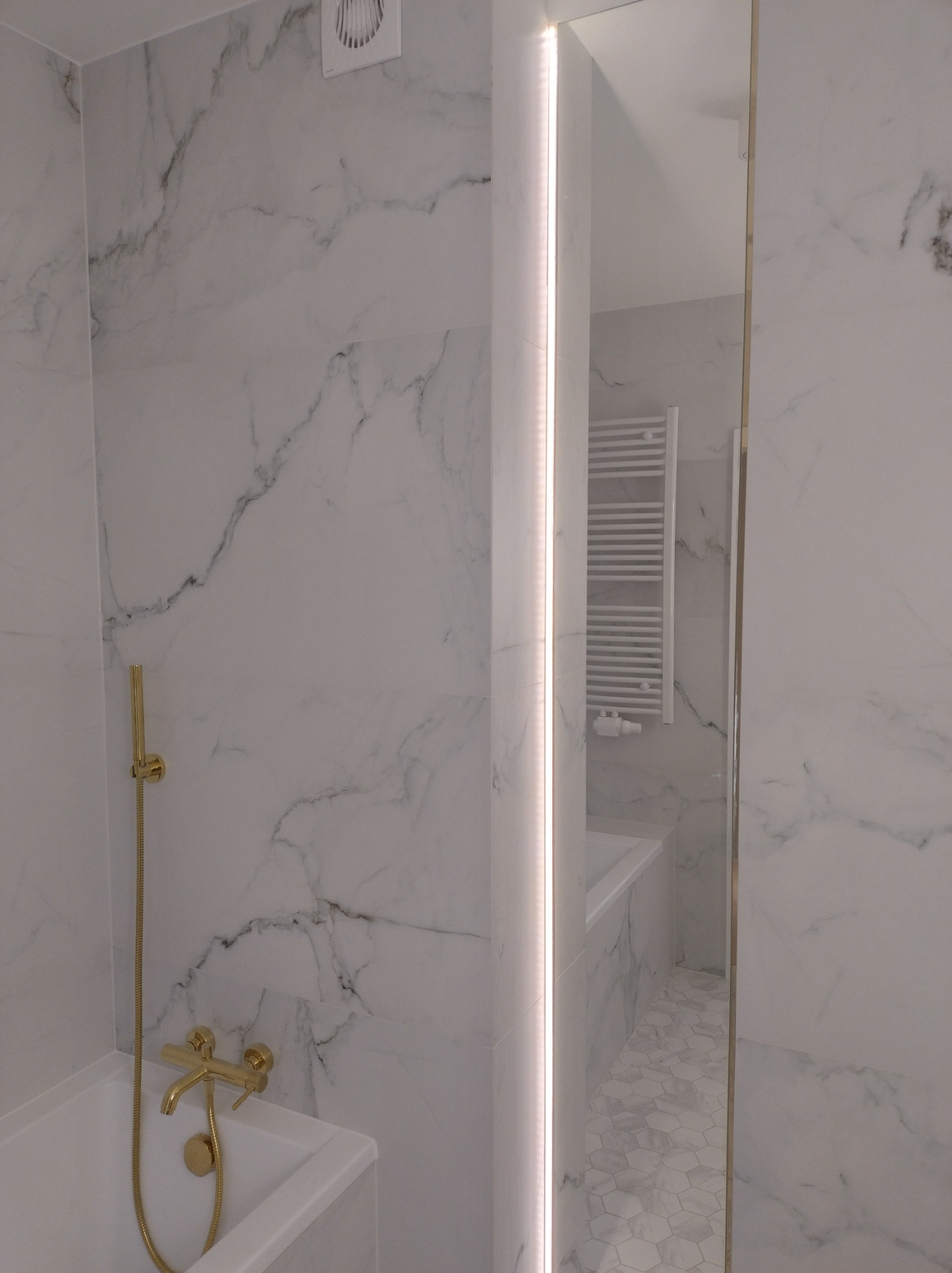 Wykończona łazienka z białymi marmurowymi płytkami, złotą armaturą i heksagonalną mozaiką na podłodze, widoczny grzejnik drabinkowy i oświetlenie LED w lustrze.