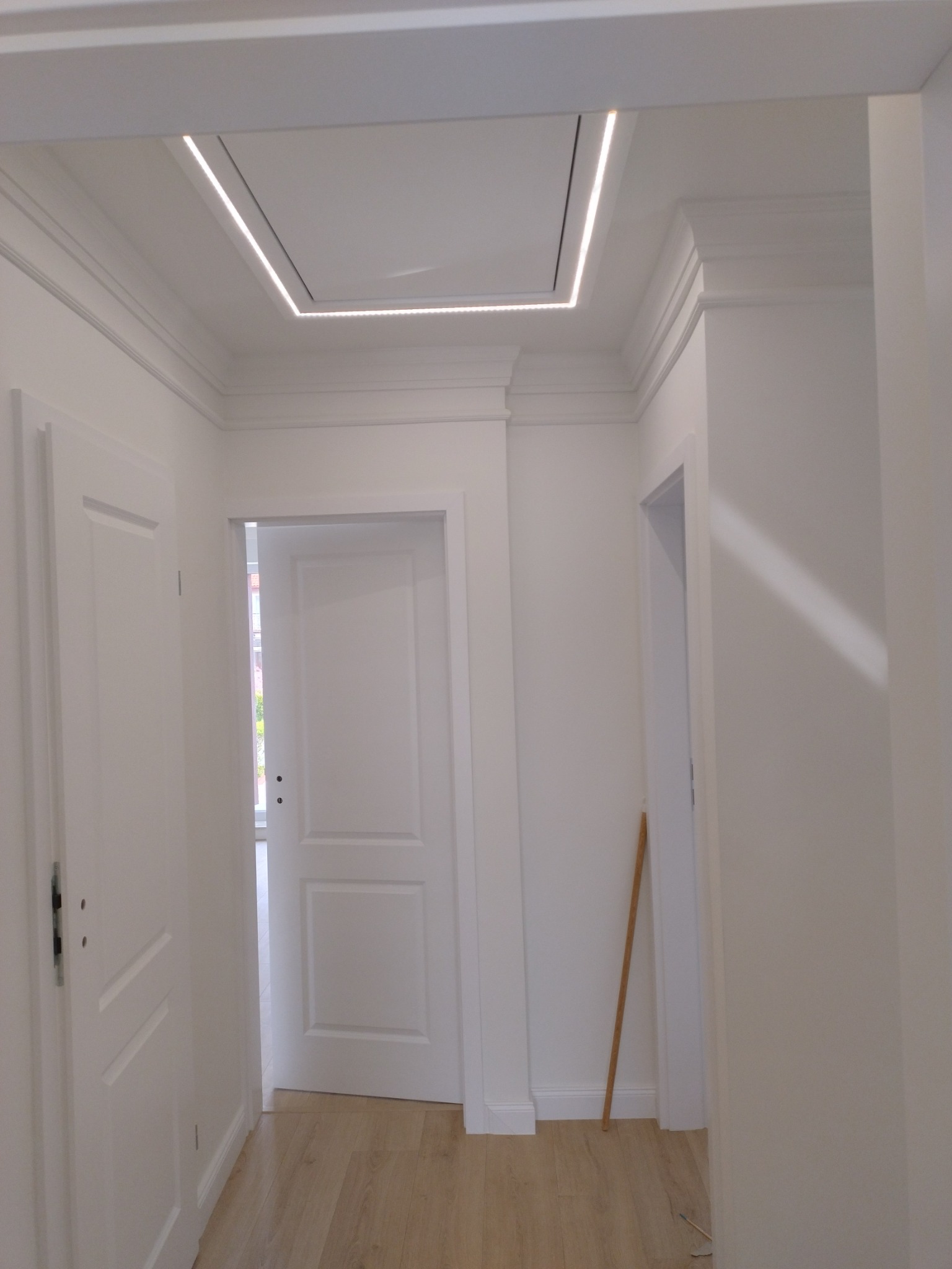 Białe wnętrze korytarza z dwiema parami drzwi i podwieszanym sufitem z oświetleniem LED w kształcie kwadratu.