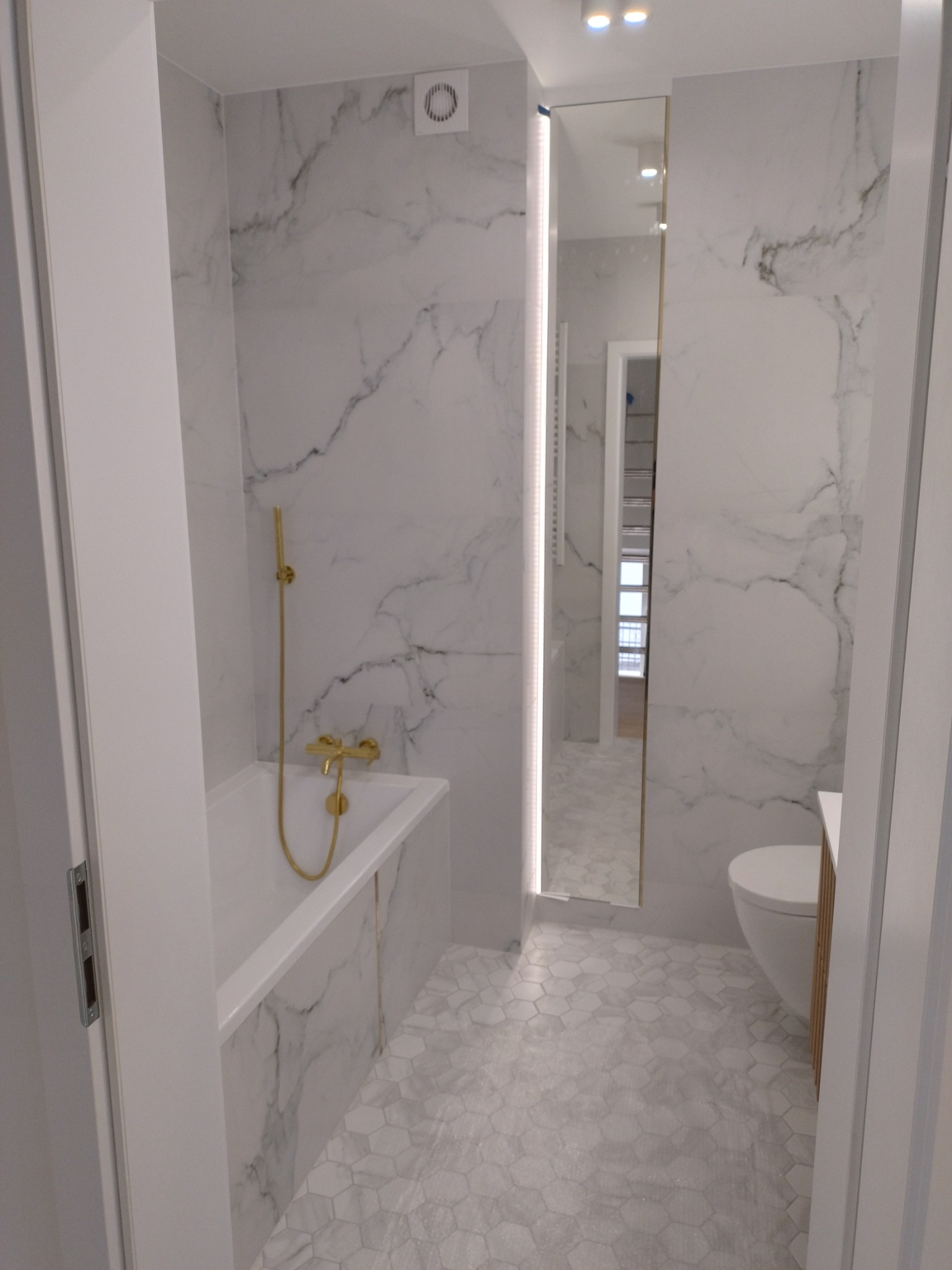 Wykończona łazienka z białymi marmurowymi płytkami na ścianach i podłodze w kształcie sześciokątów, złota armatura, lustro z podświetleniem LED, widok zza otwartych drzwi.