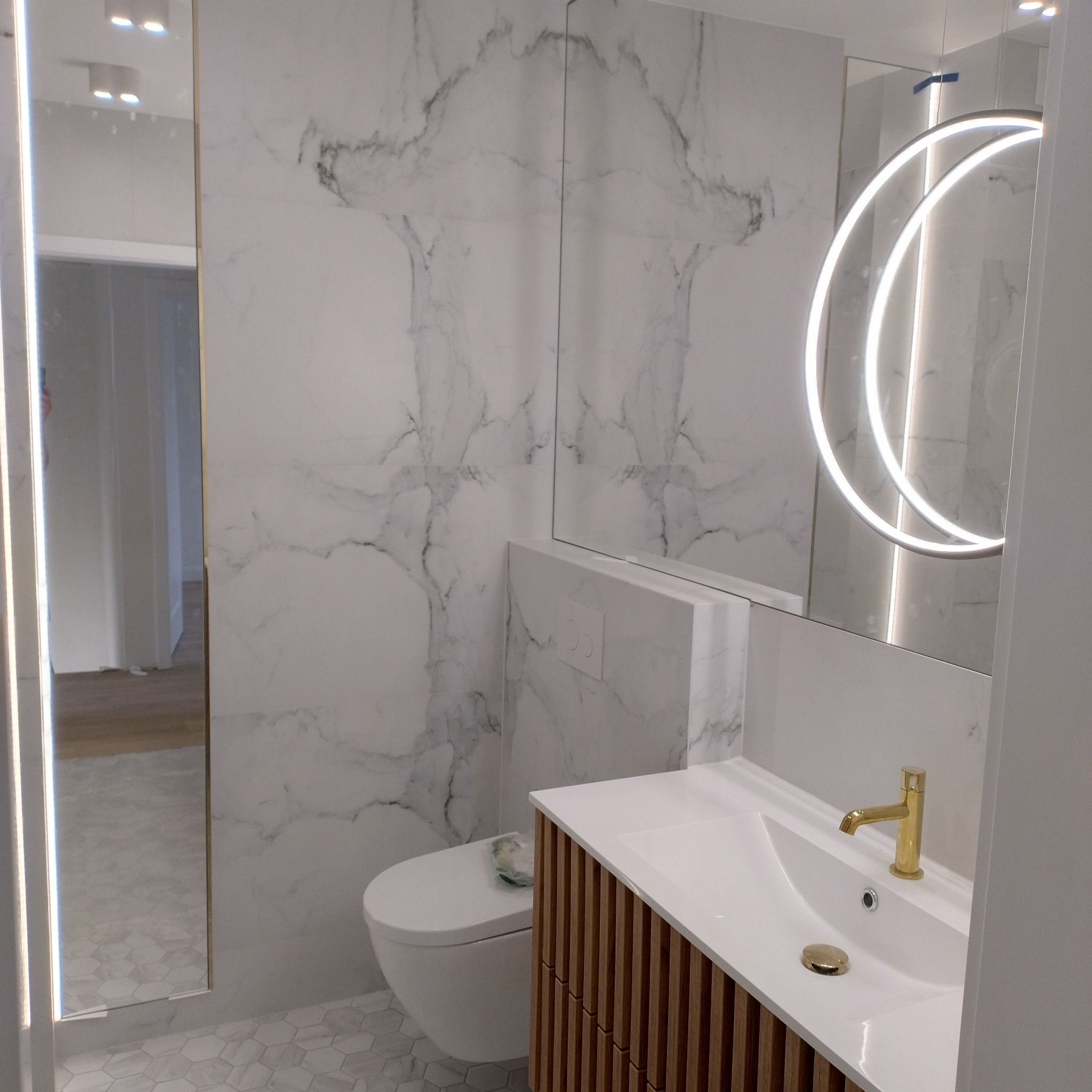 Nowoczesna łazienka z marmurowymi płytkami, podwieszaną toaletą, szafką pod umywalką z drewnianymi lamelami i okrągłym lustrem podświetlanym LED.