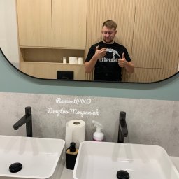 RemontPRO Usługi remontowo-wykończeniowe Dmytro Mospaniuk - Nowoczesna łazienka z dwiema białymi umywalkami nablatowymi, czarnymi bateriami i dużym owalnym lustrem, w którym odbija się mężczyzna robiący sobie zdjęcie telefonem.