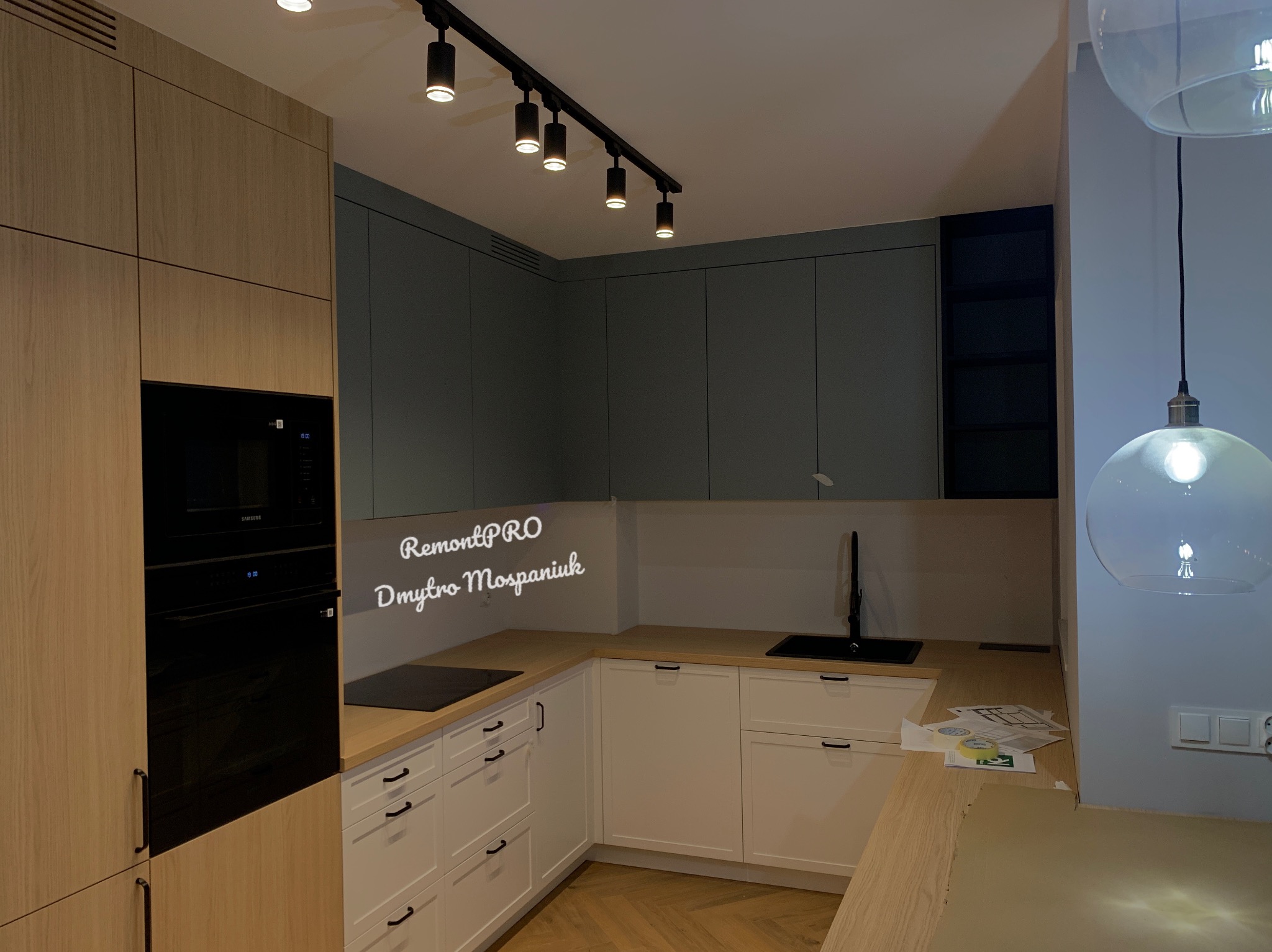 Nowoczesna kuchnia z drewnianymi i szarymi szafkami, czarnym piekarnikiem i płytą grzewczą, oświetleniem punktowym i drewnianą podłogą w jodełkę.