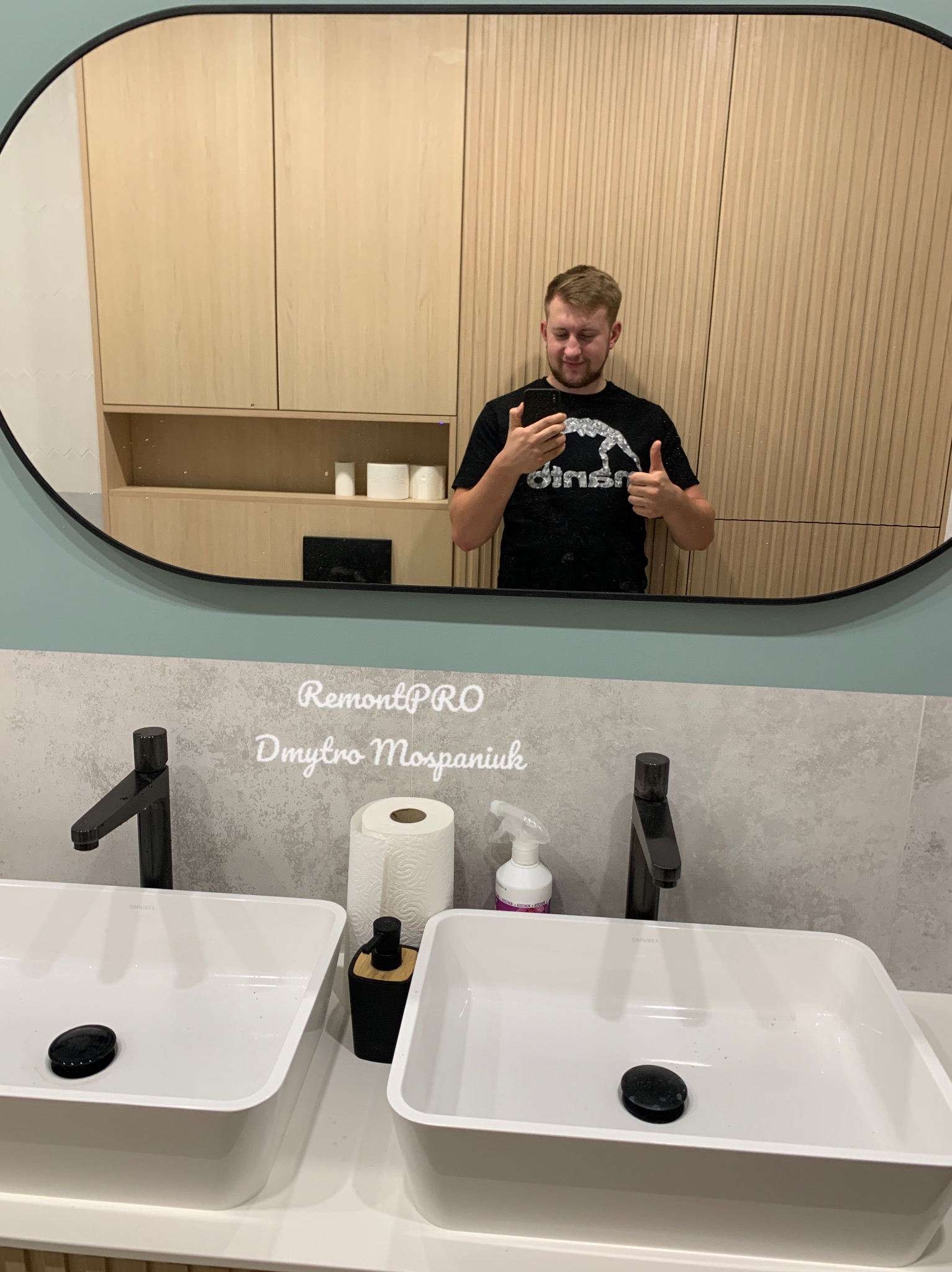 Nowoczesna łazienka z dwiema białymi umywalkami nablatowymi, czarnymi bateriami i dużym owalnym lustrem, w którym odbija się mężczyzna robiący sobie zdjęcie telefonem.