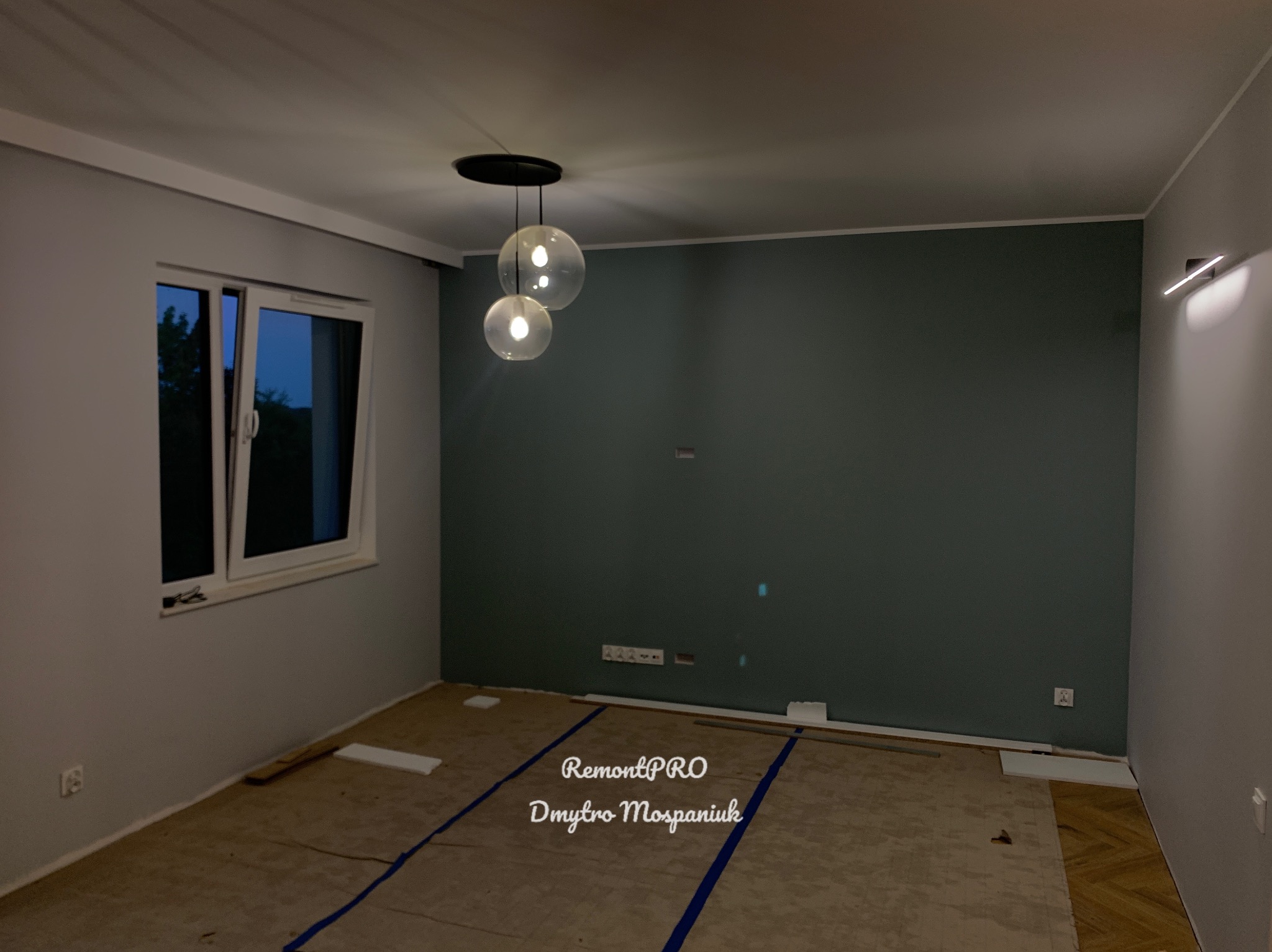 Pokój w trakcie prac wykończeniowych: pomalowane ściany w odcieniach szarości i ciemnej zieleni, widoczna instalacja oświetleniowa z kulistymi lampami, podłoga częściowo zabezpieczona przed...