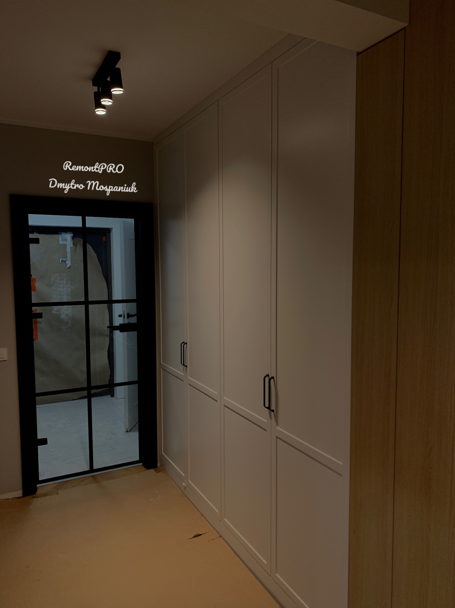 Biała zabudowa szaf wnękowych z czarnymi uchwytami, widoczna czarna rama drzwi z przeszkleniami, podłoga zabezpieczona kartonem, oświetlenie punktowe sufitowe.