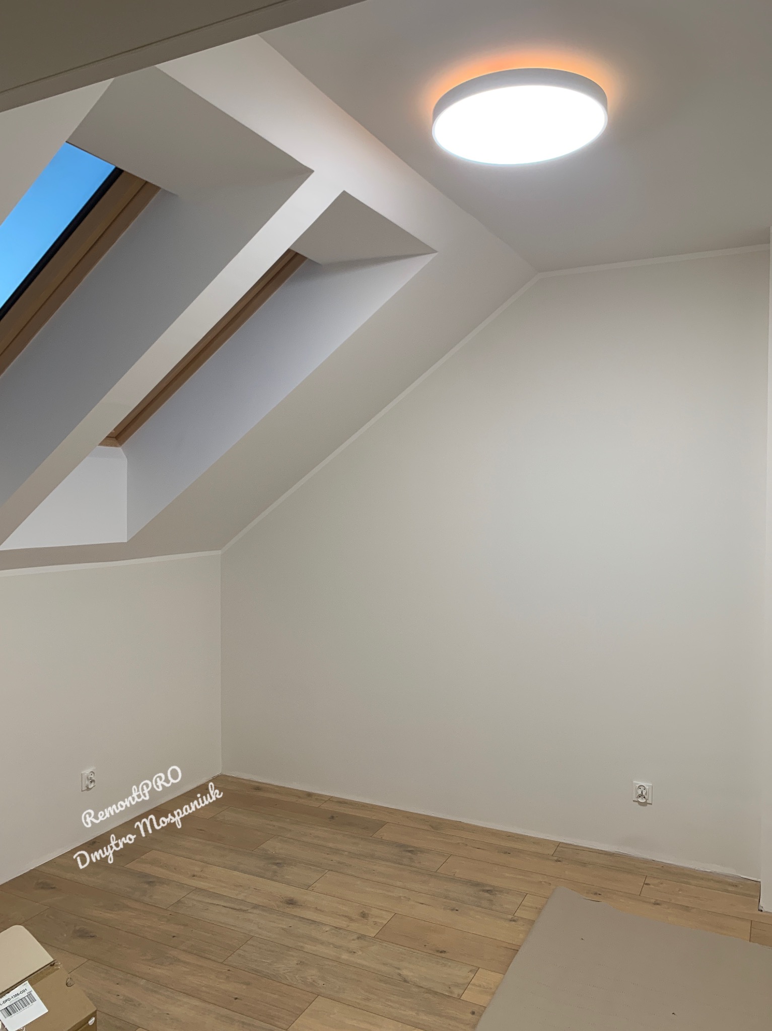 Poddasze po remoncie: jasne ściany, drewniana podłoga, okno dachowe z widokiem na niebo i nowoczesna okrągła lampa sufitowa.