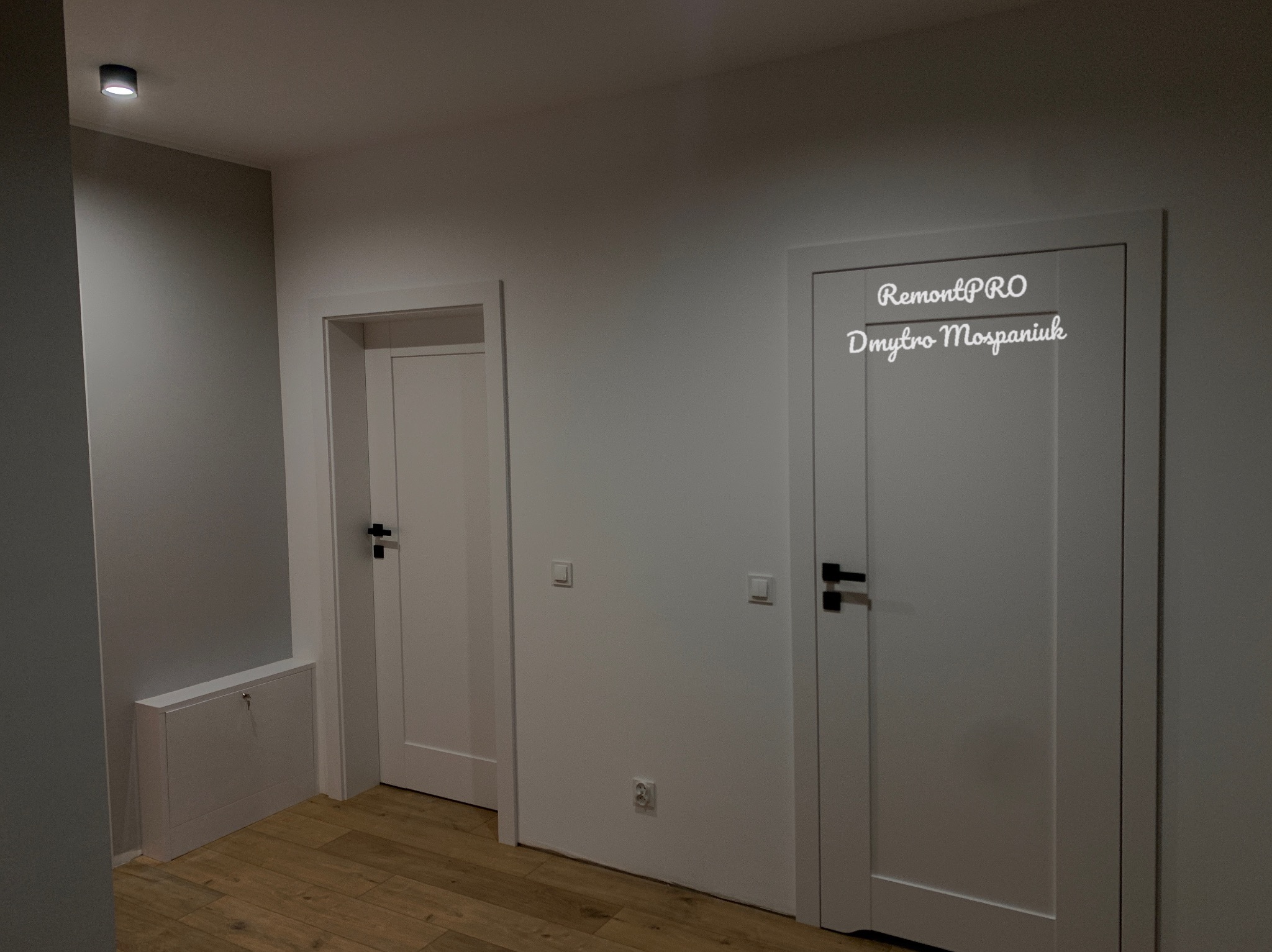 Dwa białe, wewnętrzne drzwi z czarnymi klamkami w szarej ścianie, widoczna skrzynka rewizyjna z kluczem, drewniana podłoga i logo firmy na jednym z drzwi.