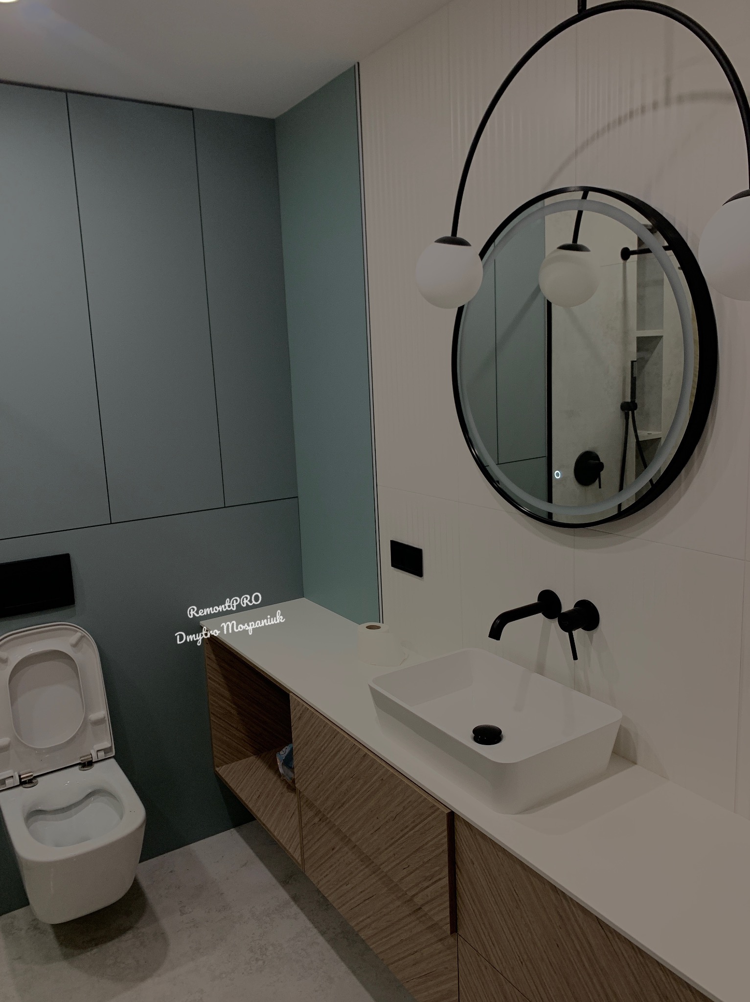 Nowoczesna łazienka z wiszącą toaletą, szafką pod umywalką z drewnianym fornirem i okrągłym lustrem z oświetleniem, minimalistyczny design.