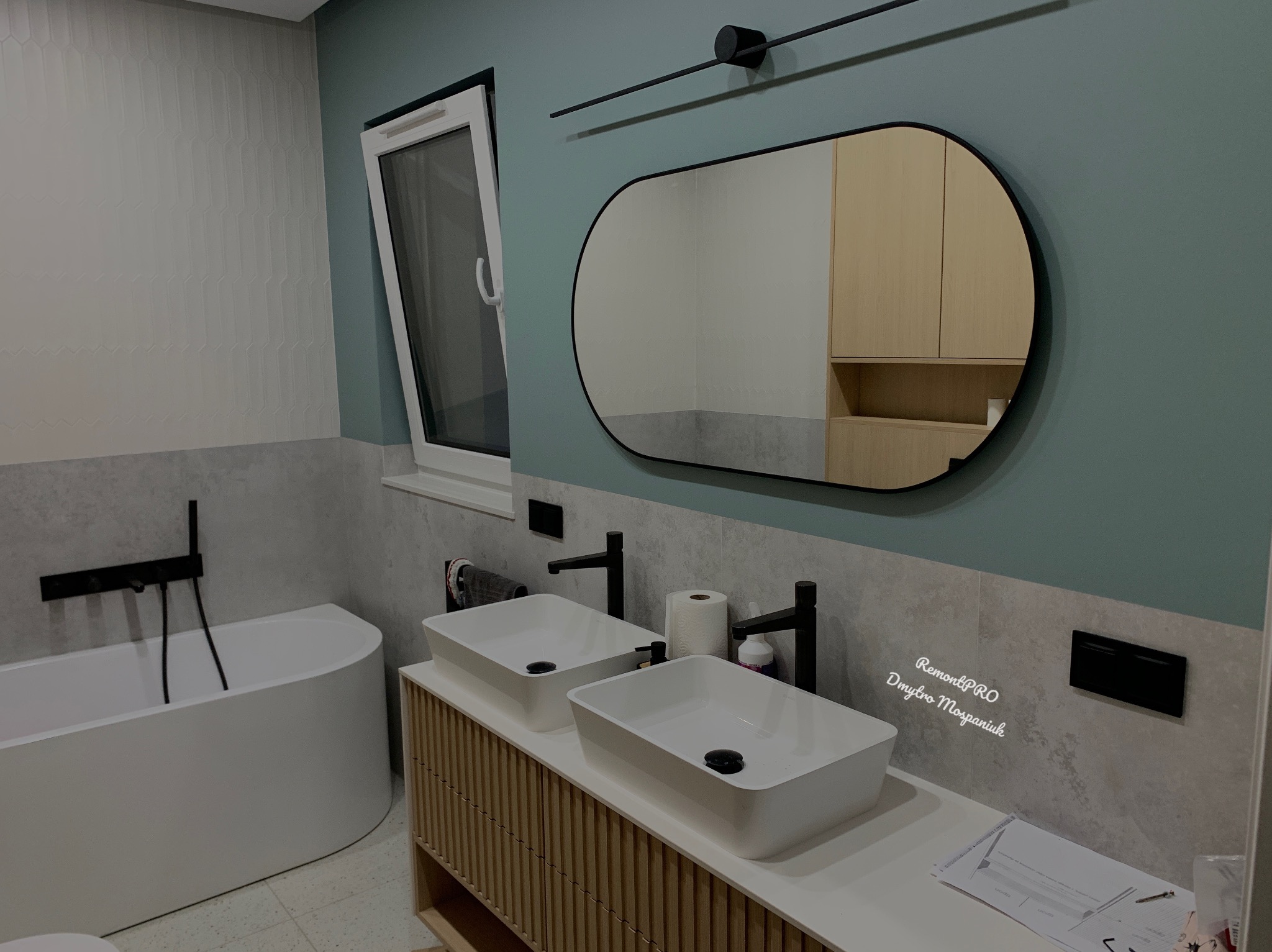 Nowoczesna łazienka z dwoma białymi umywalkami nablatowymi na szafce z lamelami, czarna armatura, owalne lustro z czarną ramą, akcenty turkusowej ściany i białe płytki w pionowy wzór, widoczna wanna.