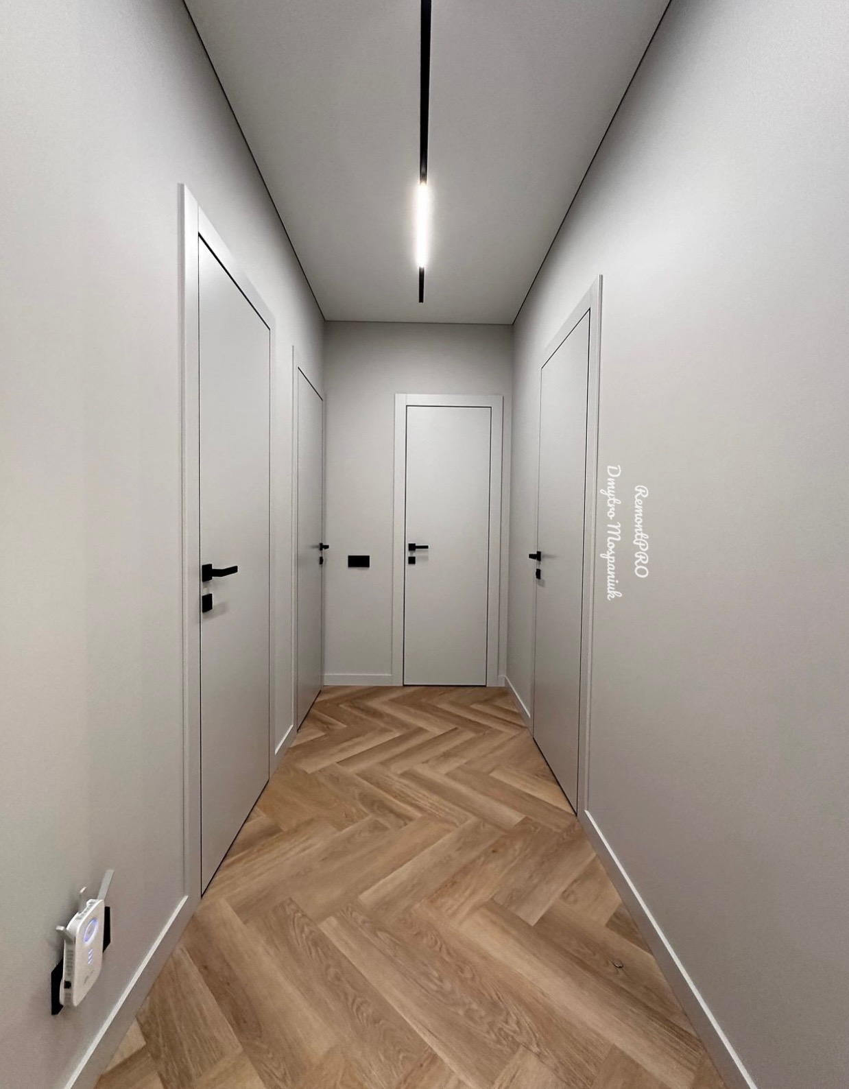 Wąski korytarz z trzema białymi drzwiami o czarnych klamkach, parkiet ułożony w jodełkę francuską, minimalistyczne oświetlenie liniowe na suficie, szare ściany i białe listwy przypodłogowe...
