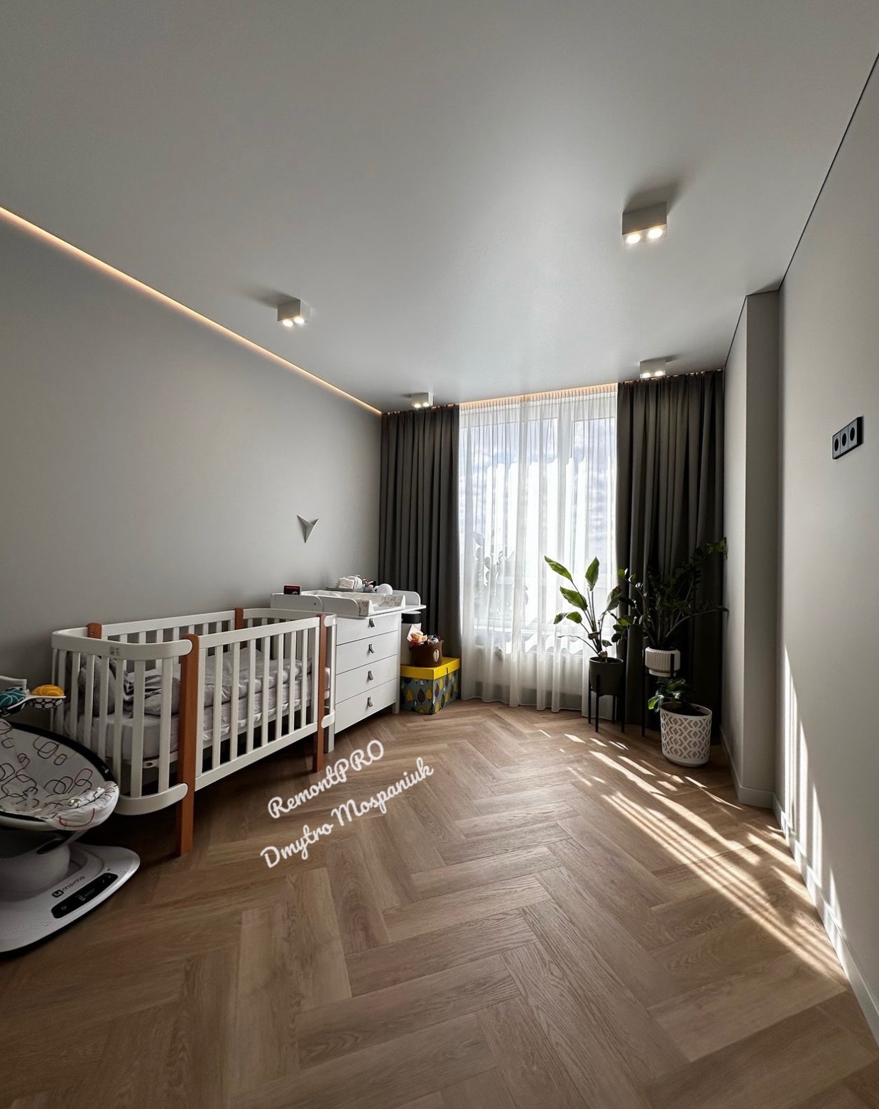 Pokój dziecięcy z białym łóżeczkiem, komodą z przewijakiem i drewnianą podłogą ułożoną w jodełkę, oświetlony światłem dziennym wpadającym przez okno z szarymi zasłonami.