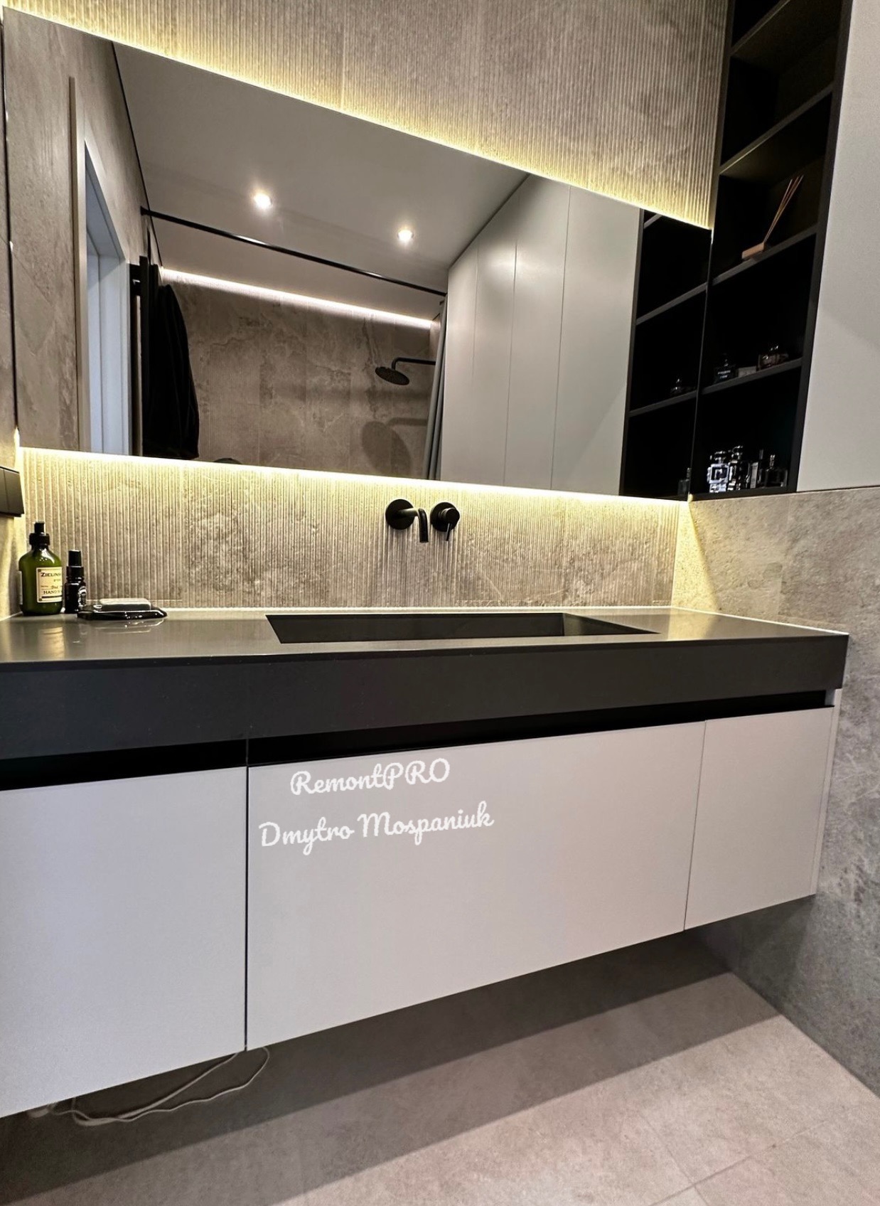 Nowoczesna łazienka z podświetlanym lustrem, czarną armaturą i szarą płytką na ścianie, widoczna zabudowa umywalkowa z białymi frontami i czarnym blatem.