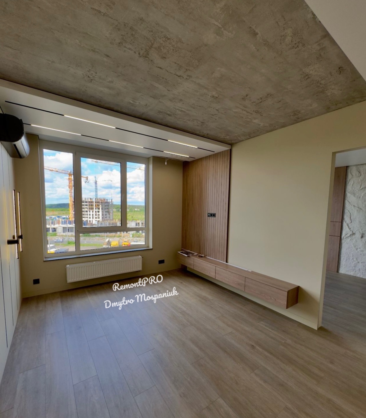 Nowoczesne wnętrze pokoju z betonowym sufitem, drewnianą podłogą, oknem z widokiem na budowę i minimalistyczną zabudową meblową.