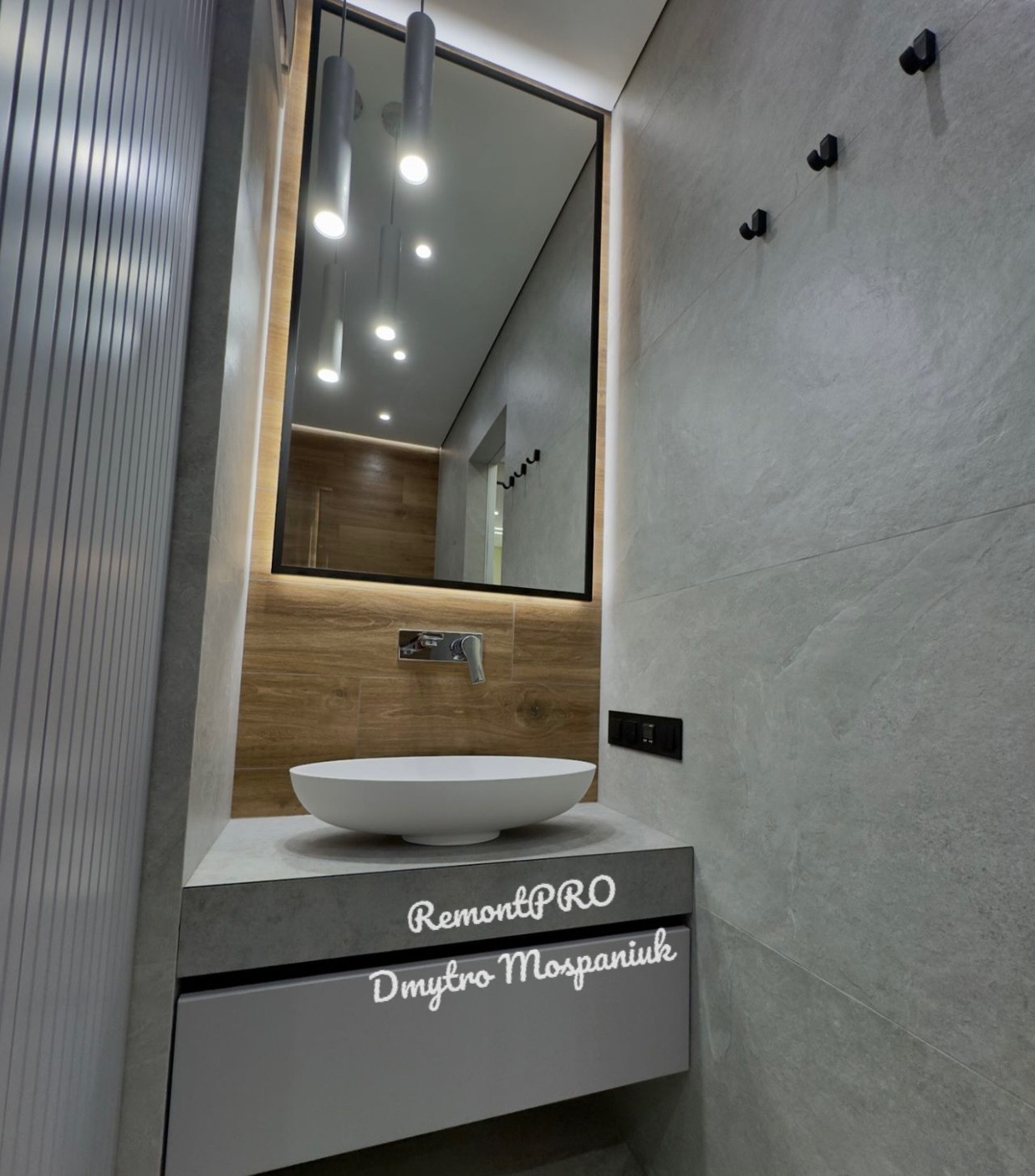 Nowoczesna łazienka z owalną umywalką nablatową, szarą szafką pod umywalką, lustrem w czarnej ramie z podświetleniem LED i szarymi płytkami imitującymi kamień na ścianach.