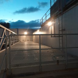 Tomasz Kubicki - Zewnętrzna platforma techniczna z metalową kratą i barierkami, oświetlona lampami, z widoczną drabiną i zbiornikami na dachu, na tle wieczornego nieba.