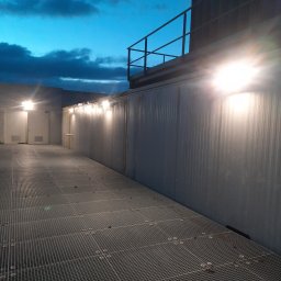 Tomasz Kubicki - Zewnętrzne oświetlenie przemysłowe na metalowej konstrukcji z kratownicą podłogową, widok nocny z zachmurzonym niebem.