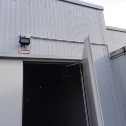 Tomasz Kubicki - Zewnętrzna ściana hali z blachy trapezowej w kolorze szarym, z zamontowanym reflektorem i rurą instalacyjną, nad otwartymi drzwiami z napisem 'Rauchabzug'.