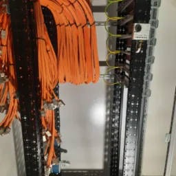 Pomarańczowe kable sieciowe uporządkowane w szafie rack, z widocznymi złączami i przewodami uziemiającymi.