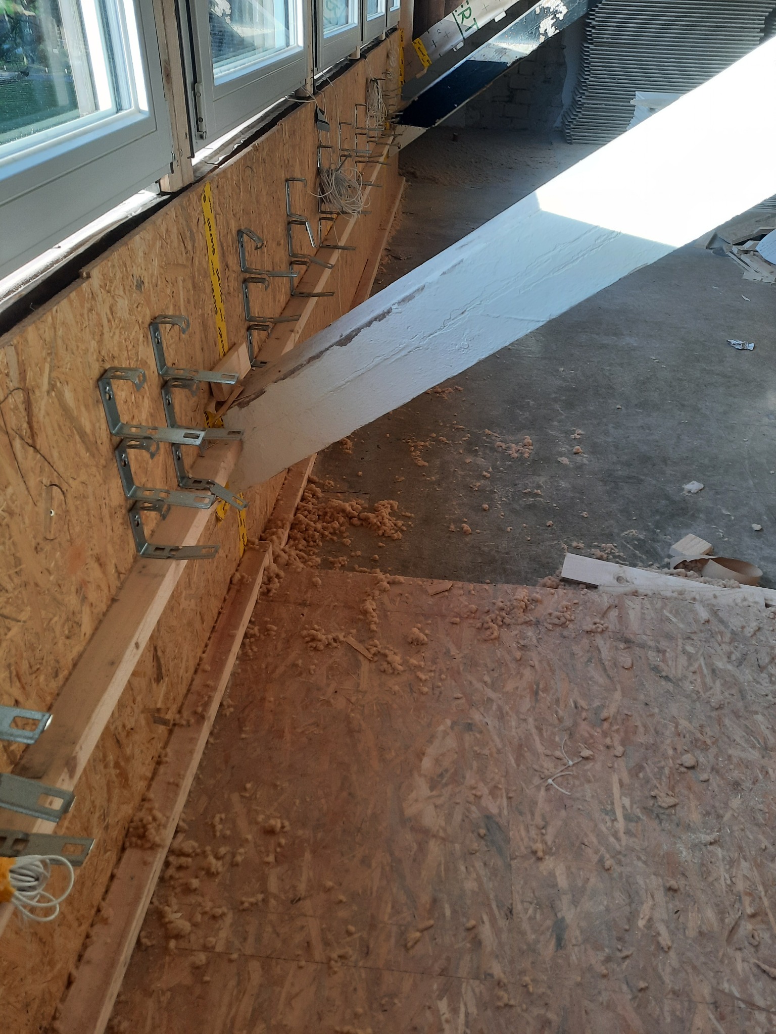 Wnętrze w trakcie prac wykończeniowych: montaż profili na ścianie z płyty OSB pod oknami, widoczne ślady pianki montażowej na podłodze i ścianie, częściowo oświetlone silnym światłem słonecznym.