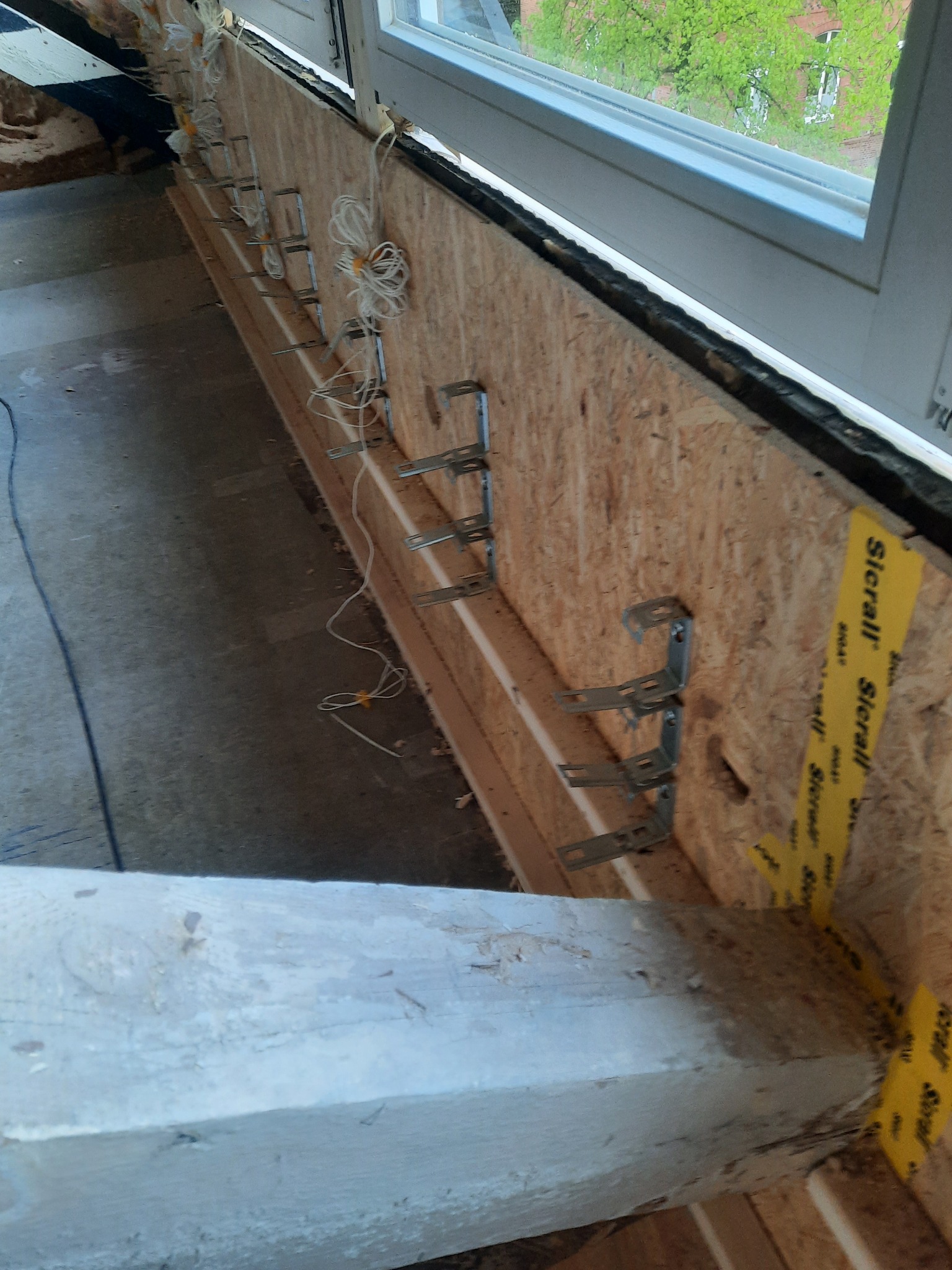 Wstępny etap montażu szyny oświetleniowej na płycie OSB pod oknem, widoczne metalowe uchwyty z przewodami i żółta taśma izolacyjna.