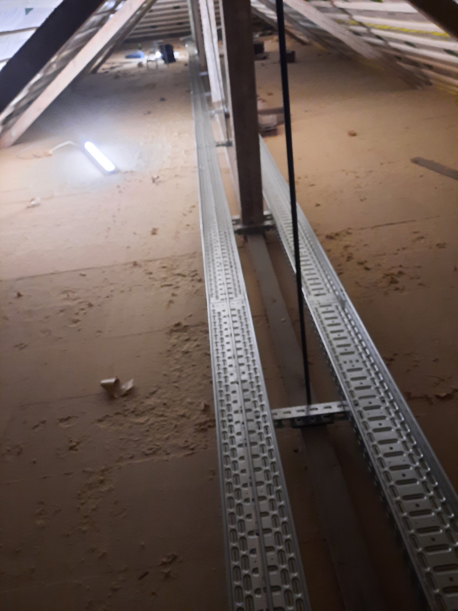 Metalowe korytka kablowe zamontowane w przestrzeni poddasza, widoczne elementy konstrukcji dachu i izolacja, oświetlenie robocze w tle.