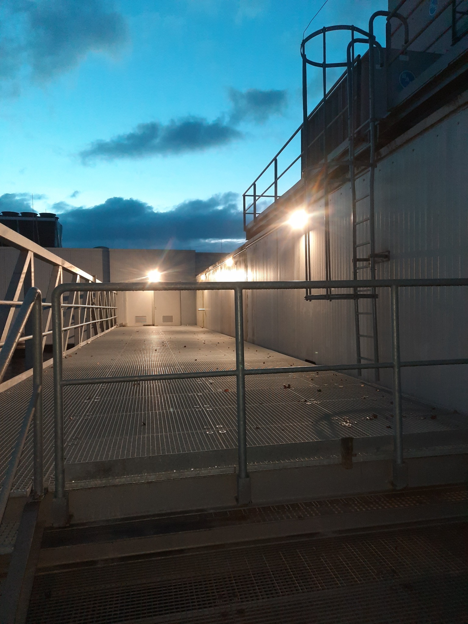 Zewnętrzna platforma techniczna z metalową kratą i barierkami, oświetlona lampami, z widoczną drabiną i zbiornikami na dachu, na tle wieczornego nieba.