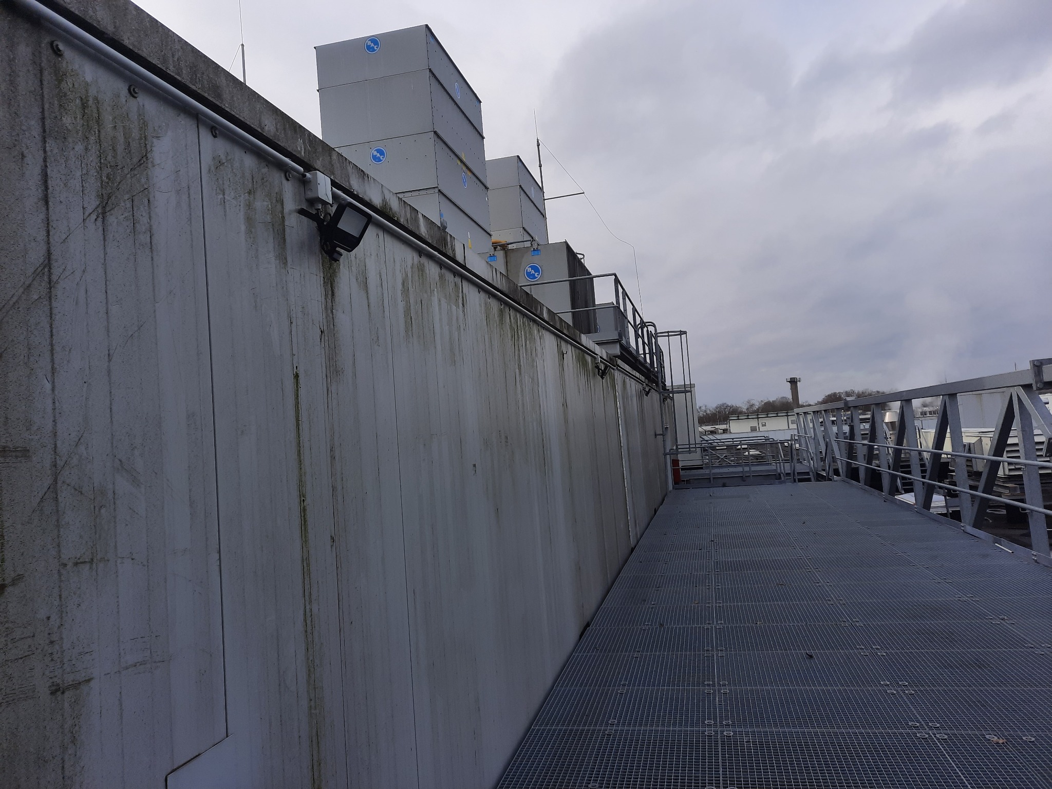 Szara, betonowa ściana budynku przemysłowego z widocznymi śladami zabrudzeń i zamontowanym oświetleniem zewnętrznym, na górze widoczne urządzenia chłodnicze z logo BAC i instalacja odgromowa,...