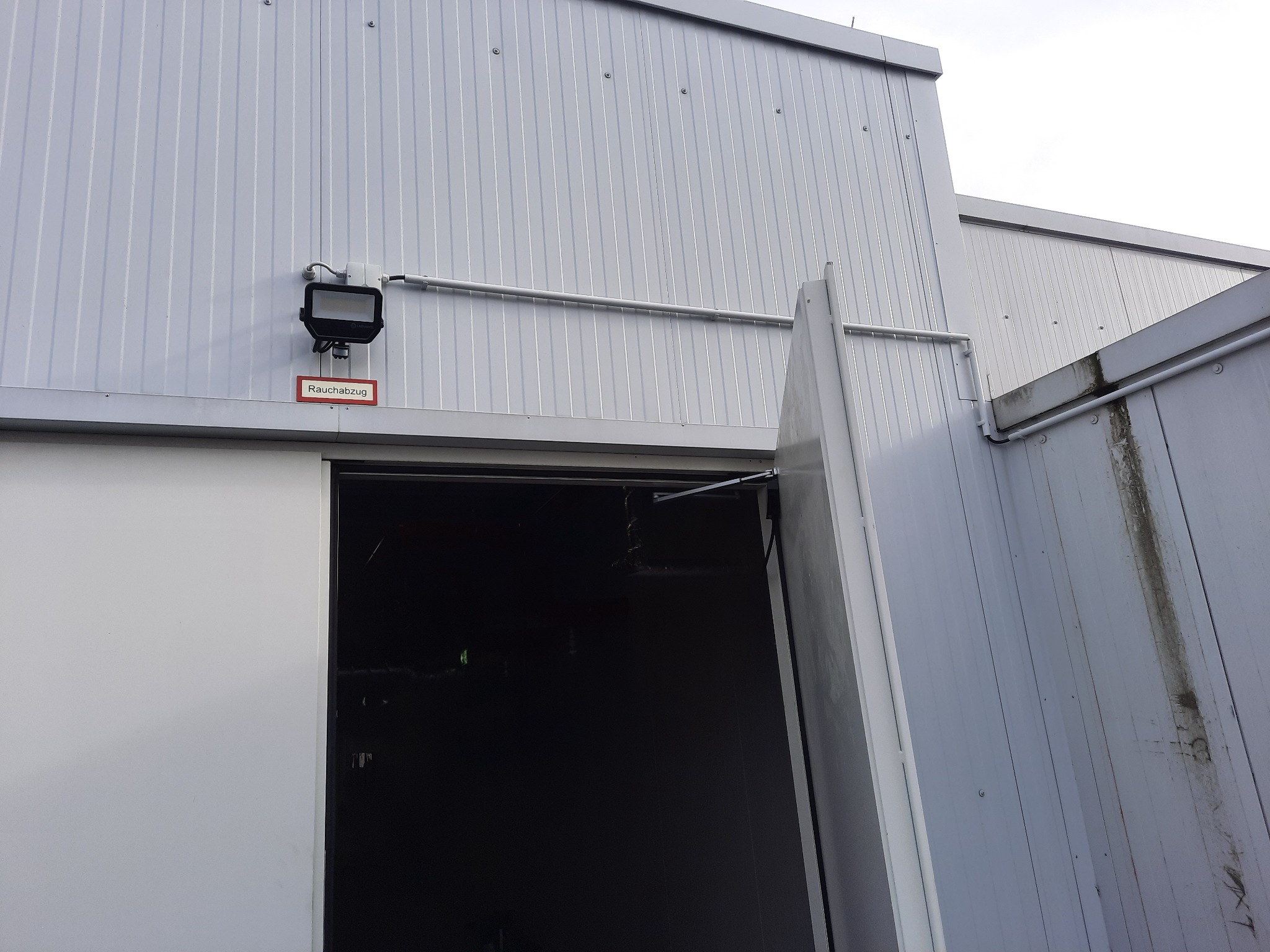 Zewnętrzna ściana hali z blachy trapezowej w kolorze szarym, z zamontowanym reflektorem i rurą instalacyjną, nad otwartymi drzwiami z napisem 'Rauchabzug'.