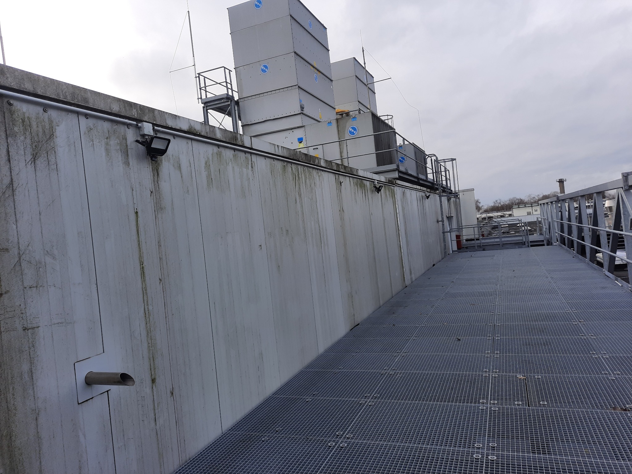 Instalacja odgromowa na dachu budynku przemysłowego z widocznymi elementami systemu, w tym metalowe maszty i przewody, oraz oświetlenie zewnętrzne, całość w szarej tonacji z pochmurnym niebem w tle.