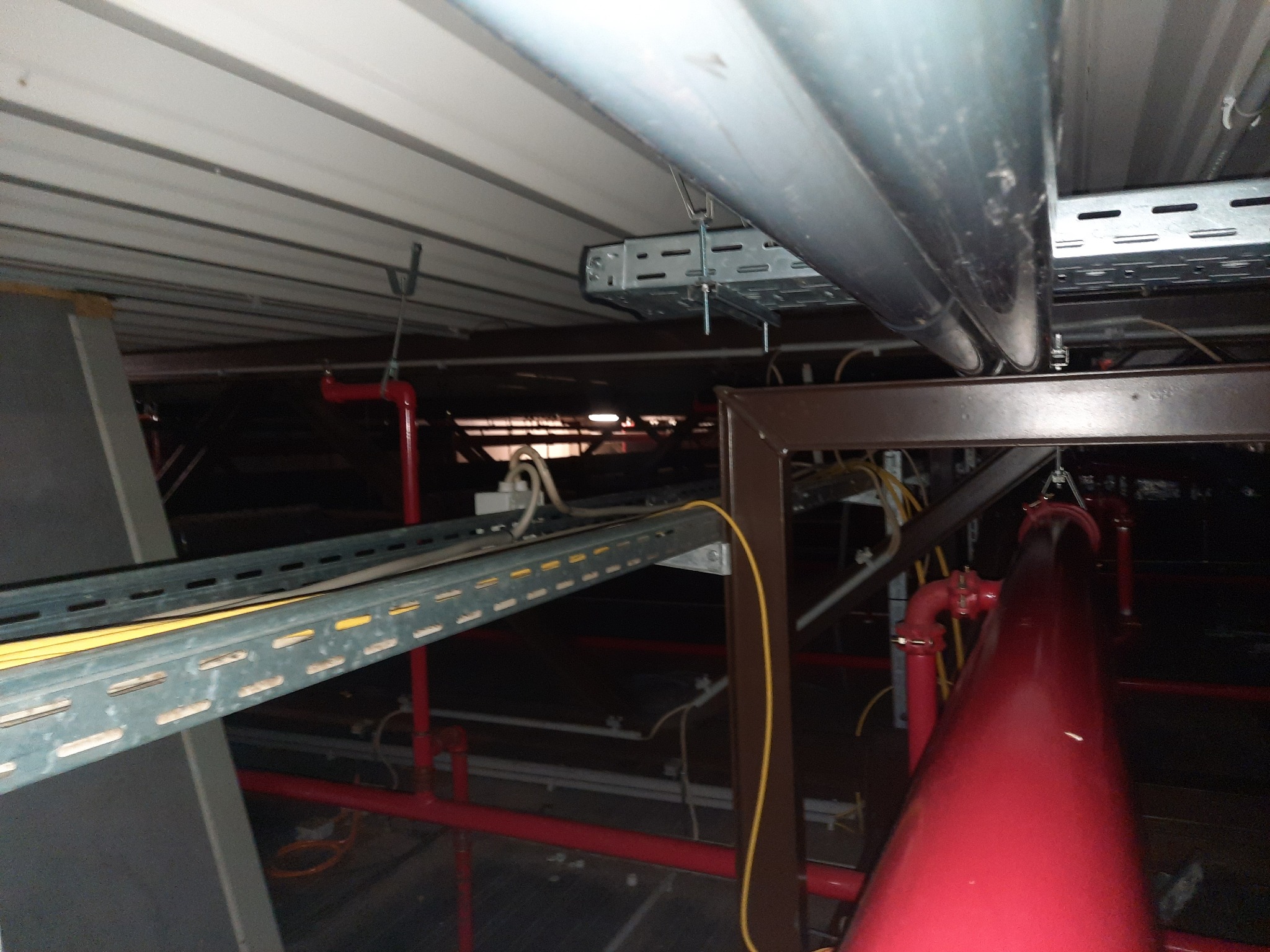 Widok instalacji pod sufitem z rurami przeciwpożarowymi w kolorze czerwonym, metalowymi korytkami kablowymi i przewodami elektrycznymi w budynku przemysłowym.