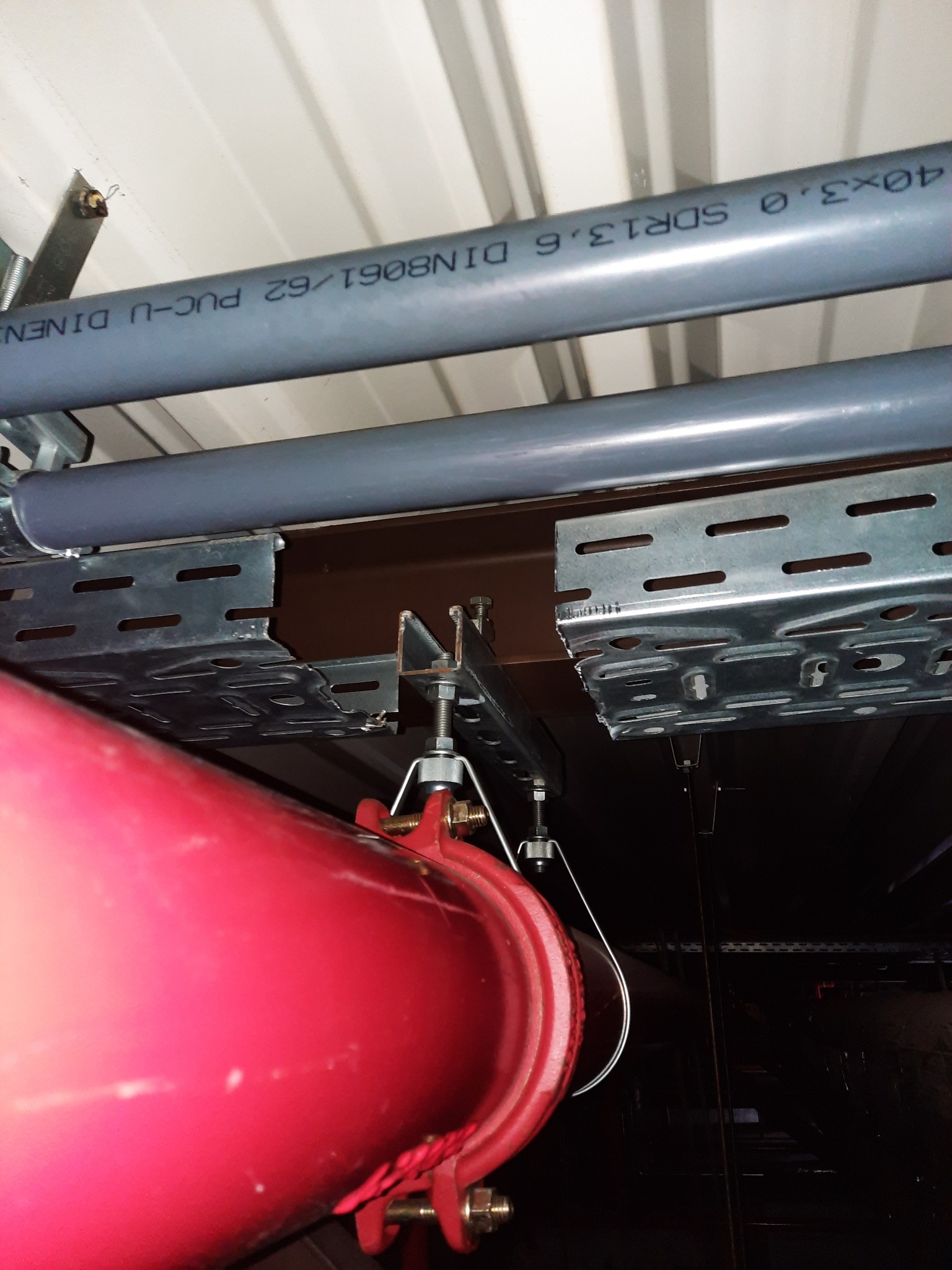 Czerwona rura instalacji tryskaczowej podwieszona pod stropem z widocznymi szarymi rurami instalacji elektrycznej i metalowymi korytkami kablowymi.