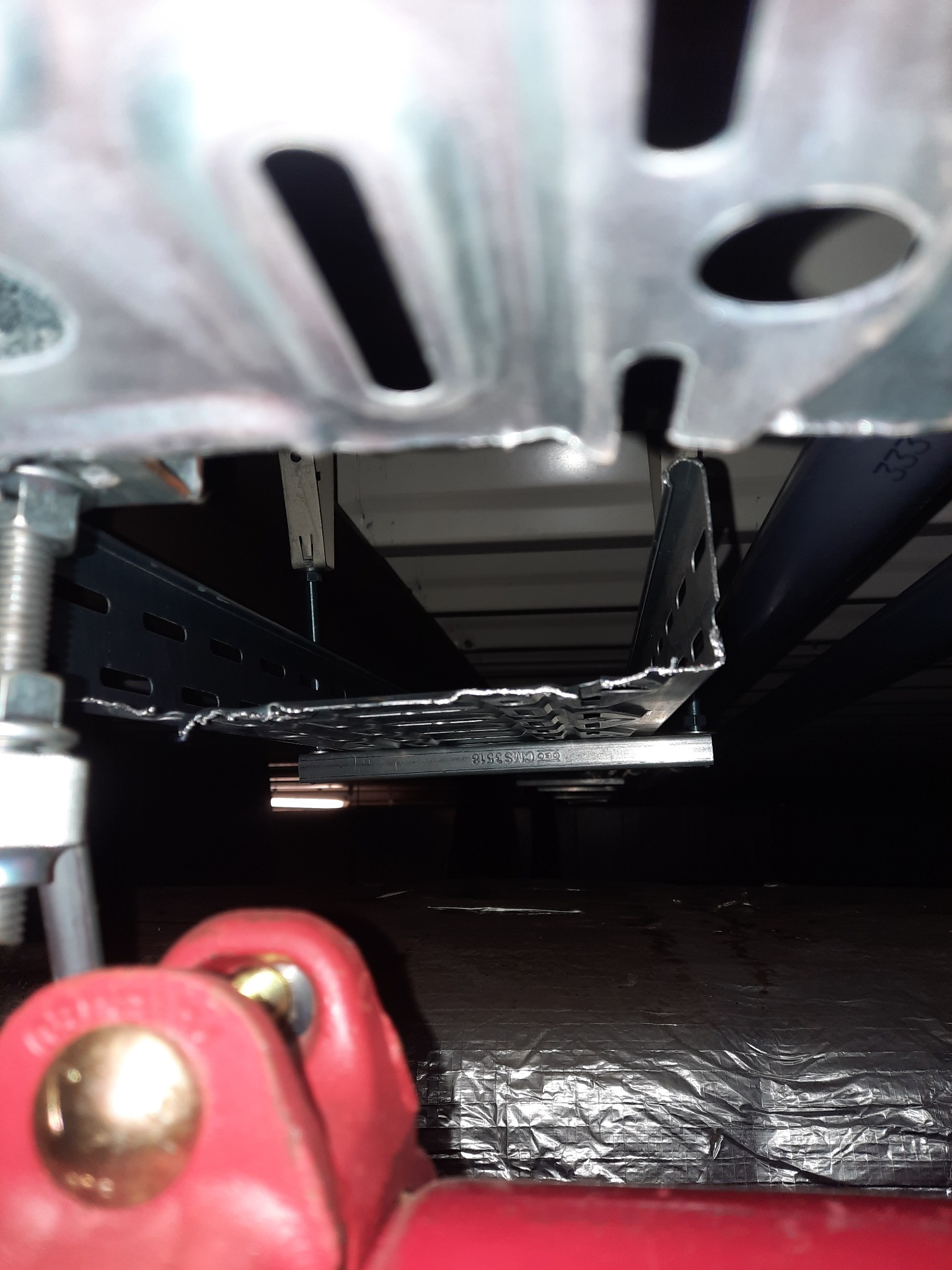 Metalowe korytka kablowe podwieszone pod sufitem, widok z dołu, z elementami mocującymi i rurami instalacyjnymi w tle.