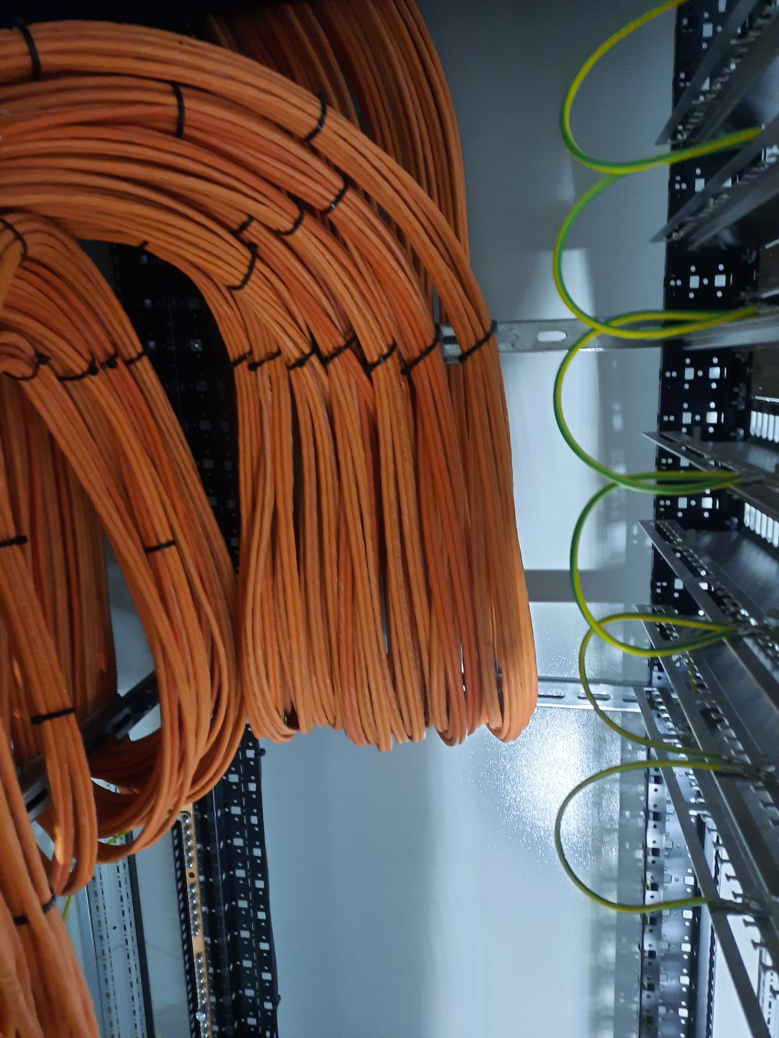 Pomarańczowe kable elektryczne ułożone w pęczki i przymocowane do metalowej konstrukcji w szafie serwerowej, widoczne również zielono-żółte przewody uziemiające.