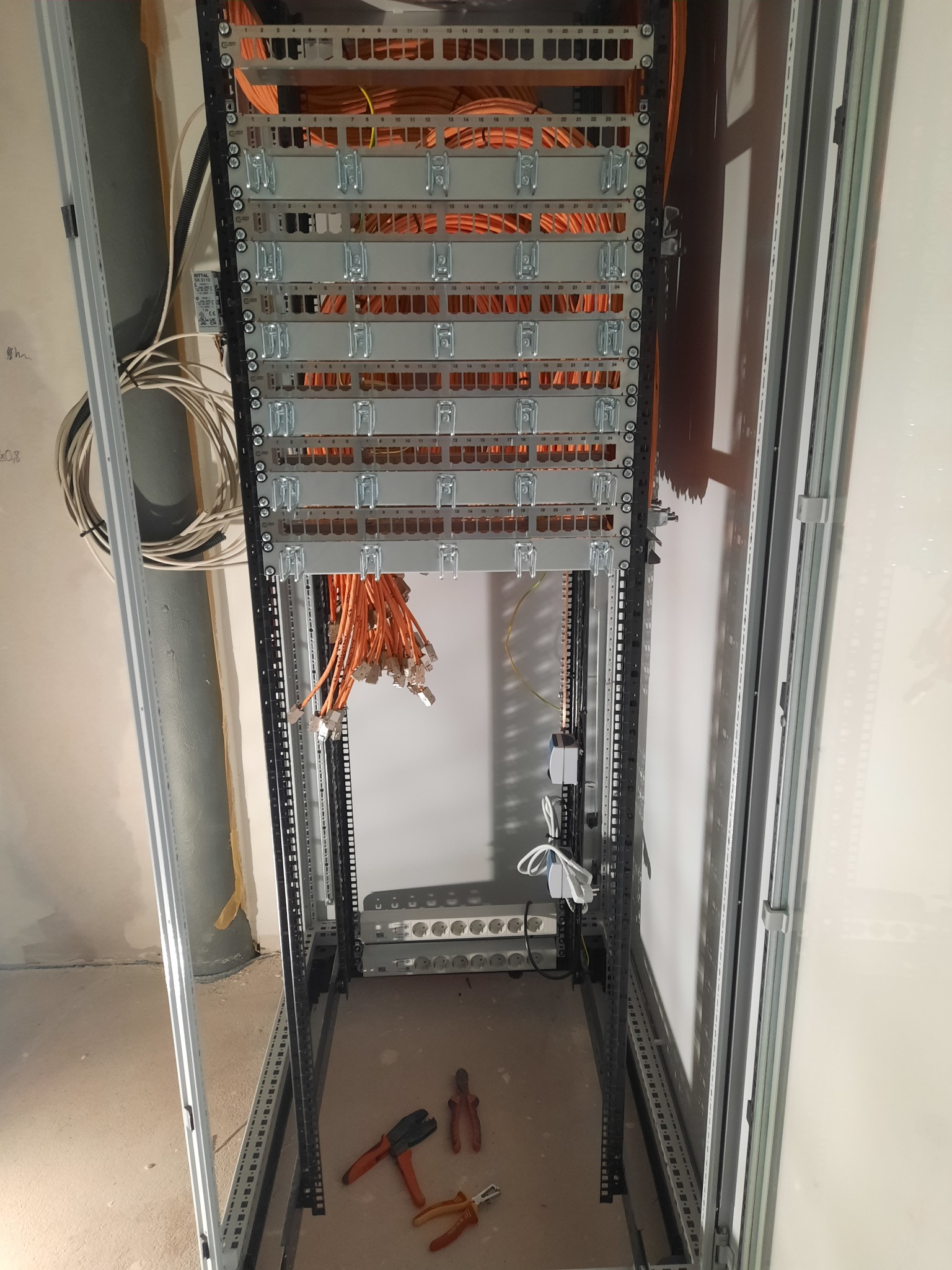 Rozdzielnica teleinformatyczna z pomarańczowymi kablami patchcord, widoczne narzędzia na podłodze, surowy stan ścian.