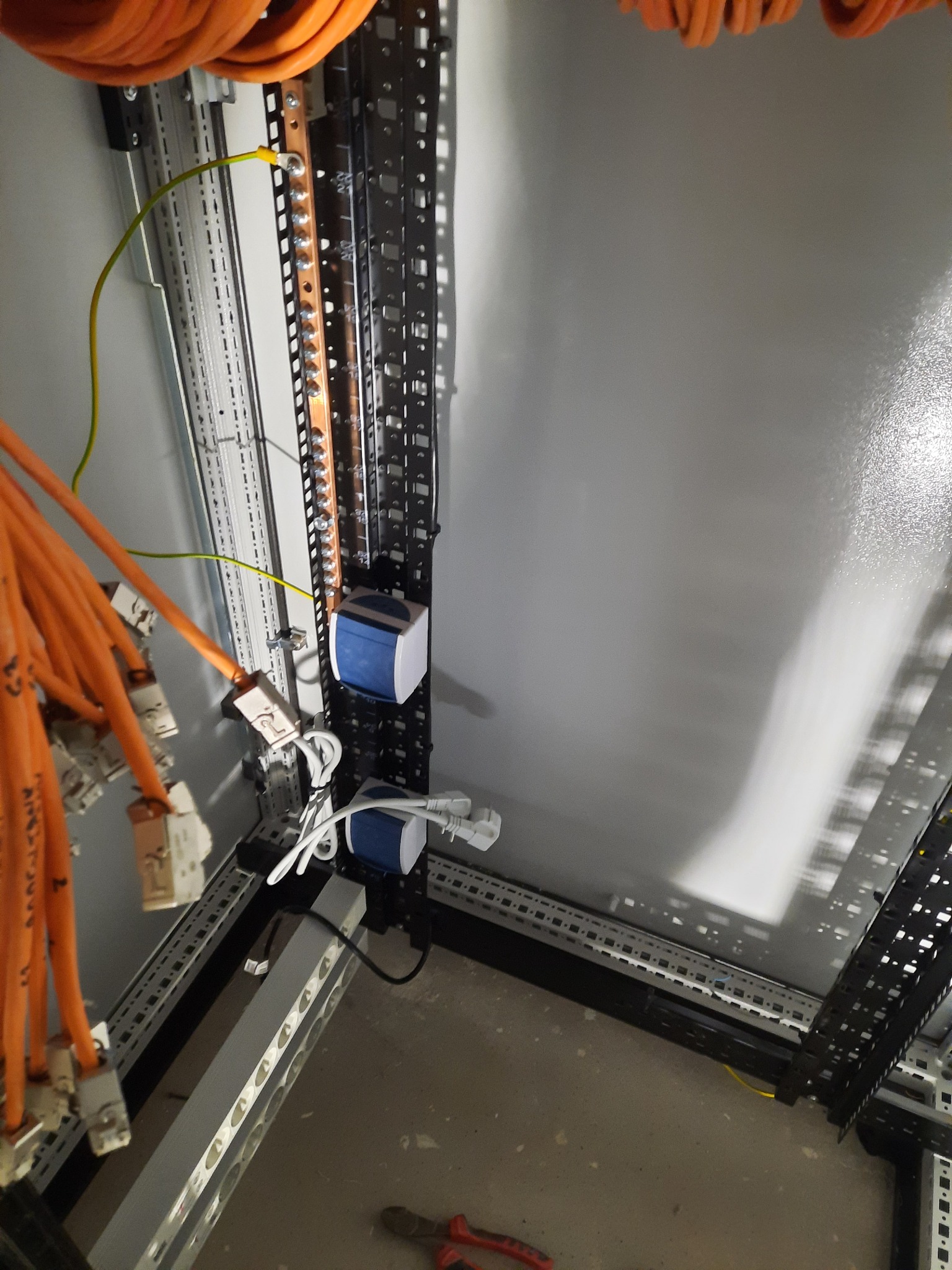 Wnętrze szafy elektrycznej w trakcie montażu, widoczne szyny DIN, przewody pomarańczowe, listwa zaciskowa z uziemieniem i obudowy zabezpieczeń, częściowo widoczne narzędzia na podłodze.