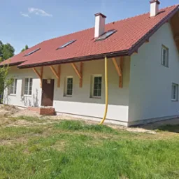 Nowo ocieplony dom z jasną elewacją i czerwoną dachówką, widoczne elementy konstrukcyjne pod dachem i żółty wąż doprowadzający materiał do ocieplenia.