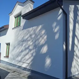 Boczny widok na świeżo pomalowaną, białą elewację domu w Radomiu, z brązowymi oknami zabezpieczonymi folią, ciemnymi rynnami i cieniem drzewa na ścianie.