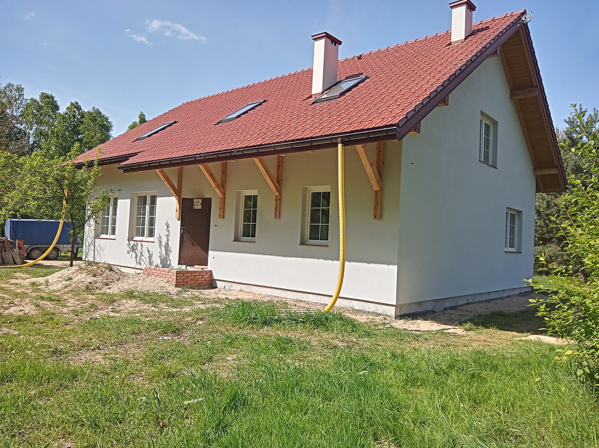 Nowo ocieplony dom z jasną elewacją i czerwoną dachówką, widoczne elementy konstrukcyjne pod dachem i żółty wąż doprowadzający materiał do ocieplenia.