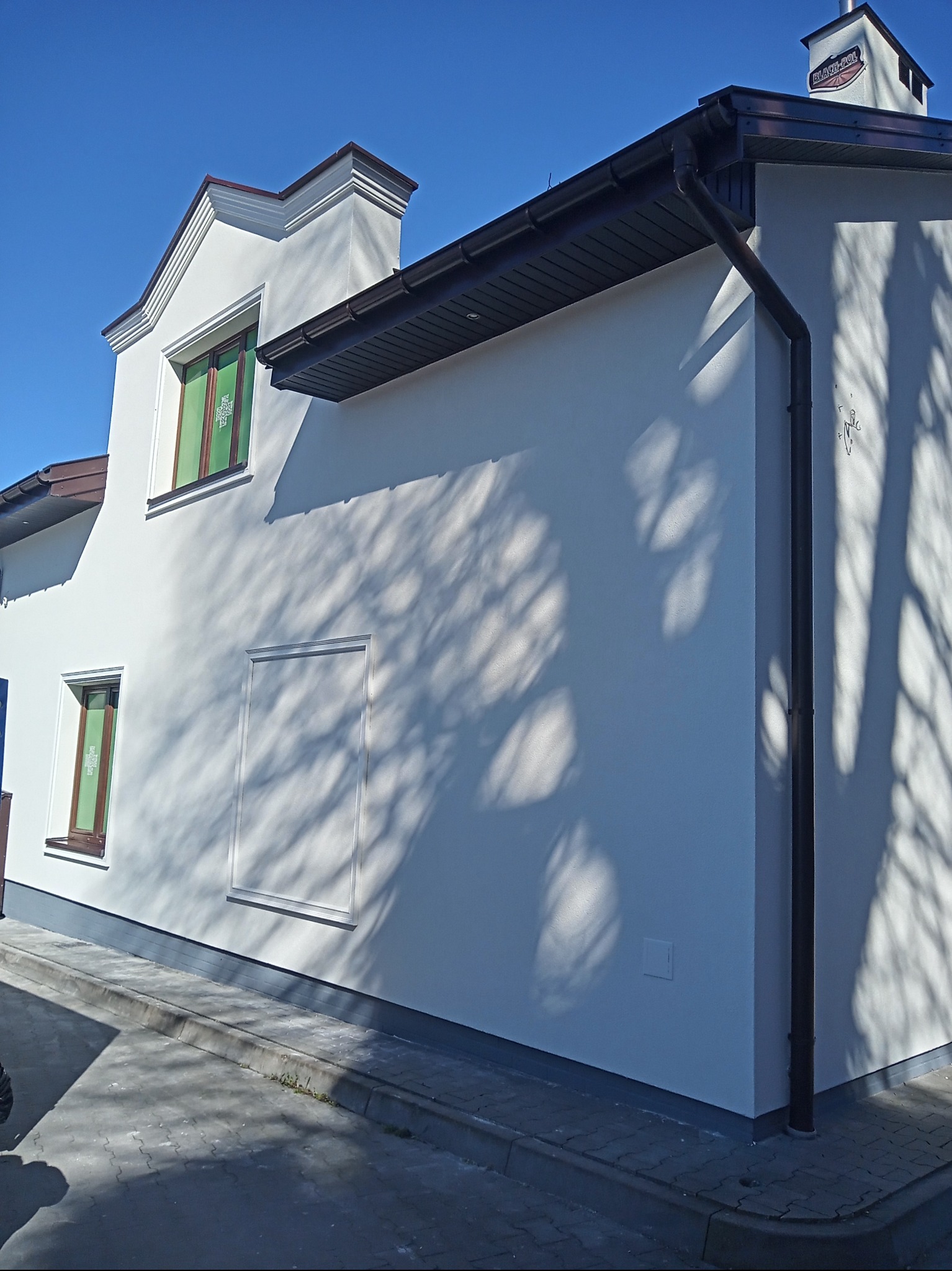 Boczny widok na świeżo pomalowaną, białą elewację domu w Radomiu, z brązowymi oknami zabezpieczonymi folią, ciemnymi rynnami i cieniem drzewa na ścianie.