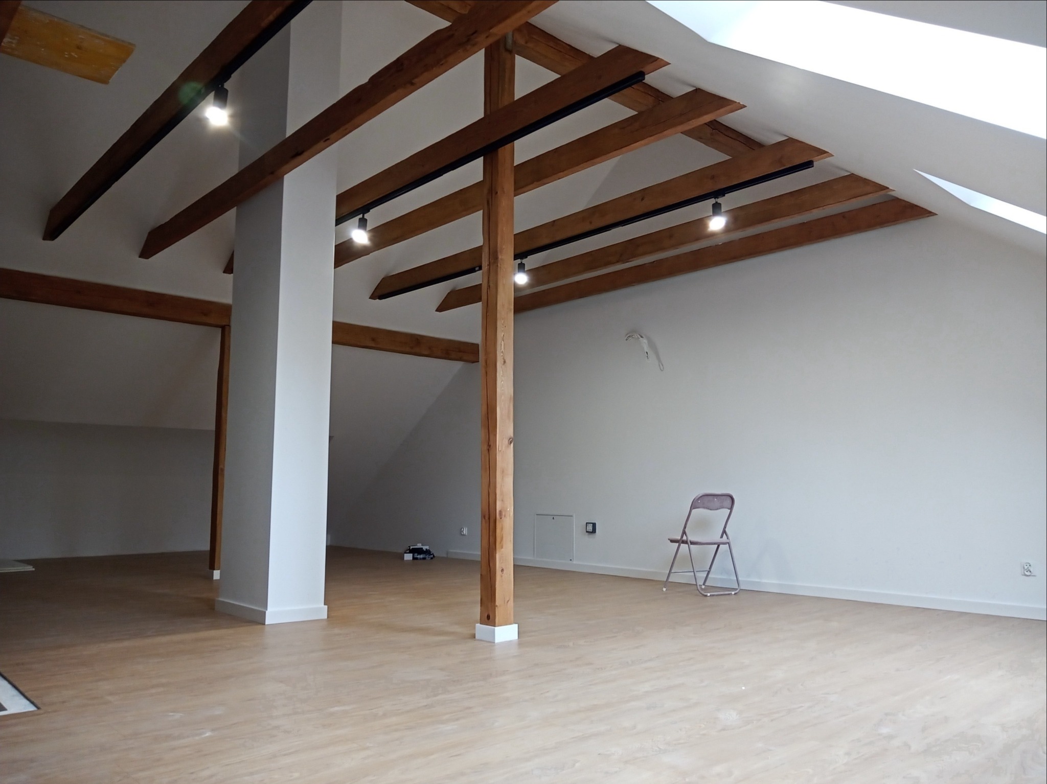 Poddasze po remoncie z drewnianymi belkami stropowymi i oświetleniem szynowym, białe ściany, jasna podłoga, pojedyncze krzesło składane