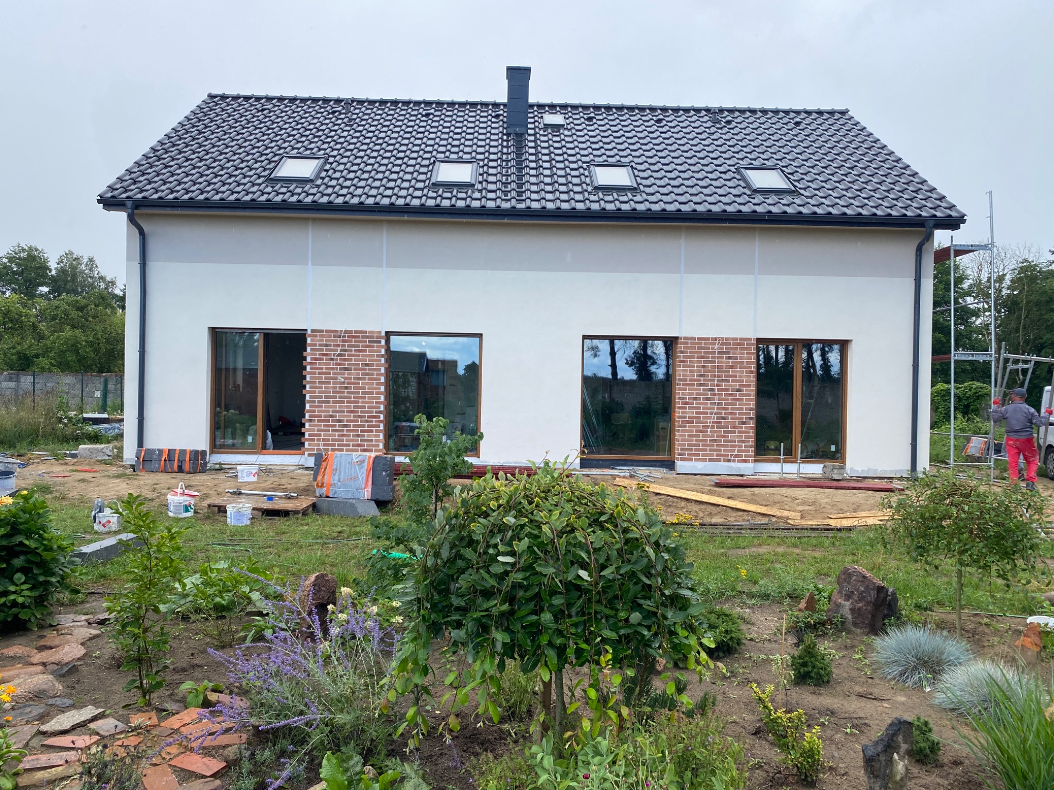 Nowoczesny dom jednorodzinny z białymi ścianami i fragmentami elewacji z czerwonej cegły, widoczne okna z drewnianą ramą, dach pokryty ciemną dachówką z trzema oknami dachowymi, teren wokół domu...