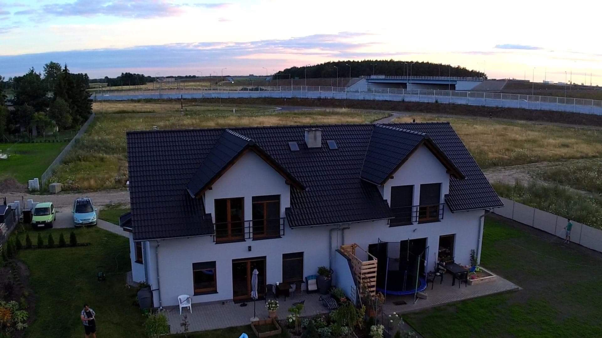 Dwupiętrowy dom z ciemnym dachem i jasną elewacją, widok z góry na ogród i okolicę z wiaduktem w tle.