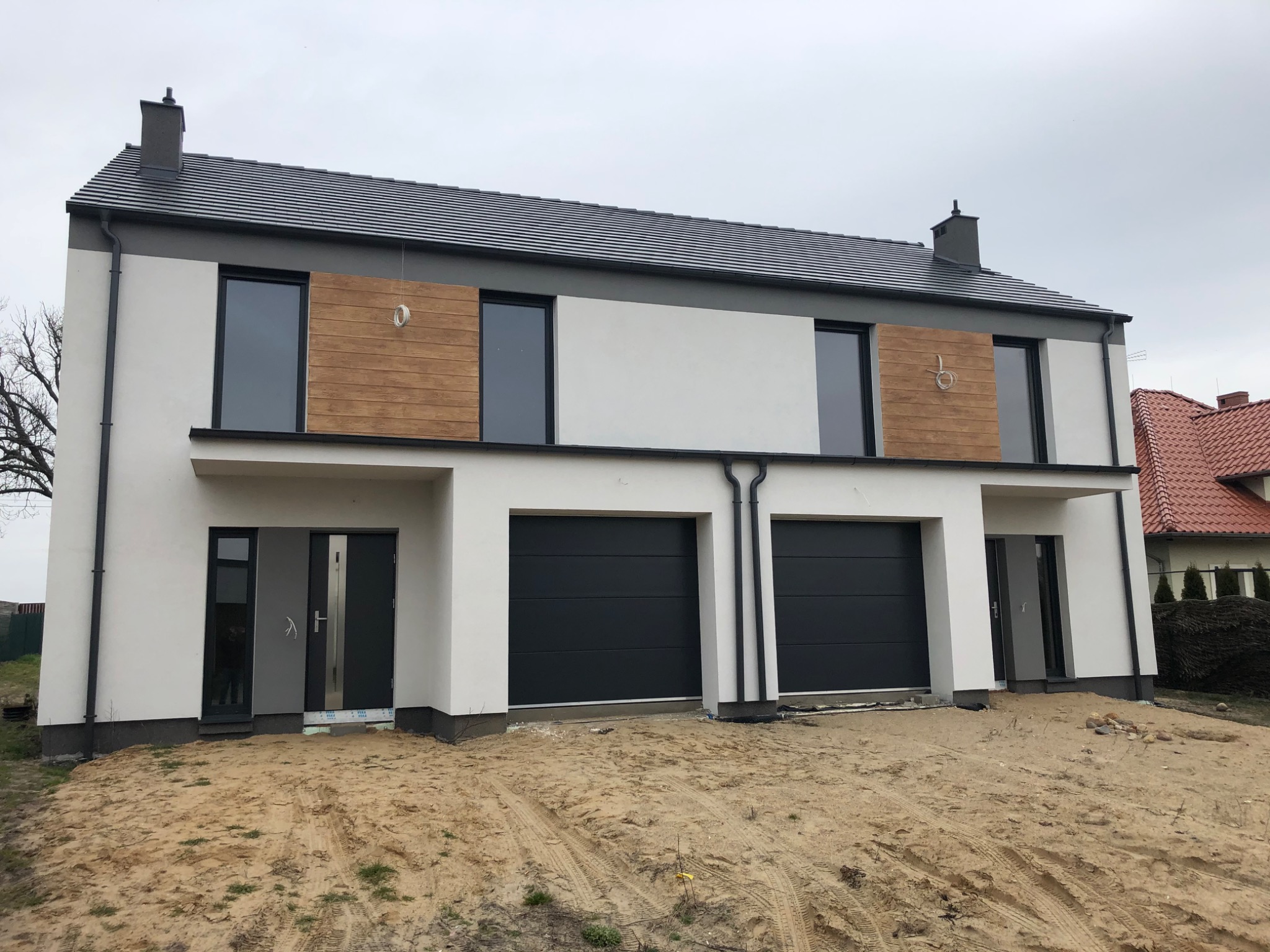 Nowoczesny bliźniak z biało-szarą elewacją, drewnianymi akcentami na piętrze i grafitowymi bramami garażowymi, teren wokół budynku w trakcie zagospodarowywania.