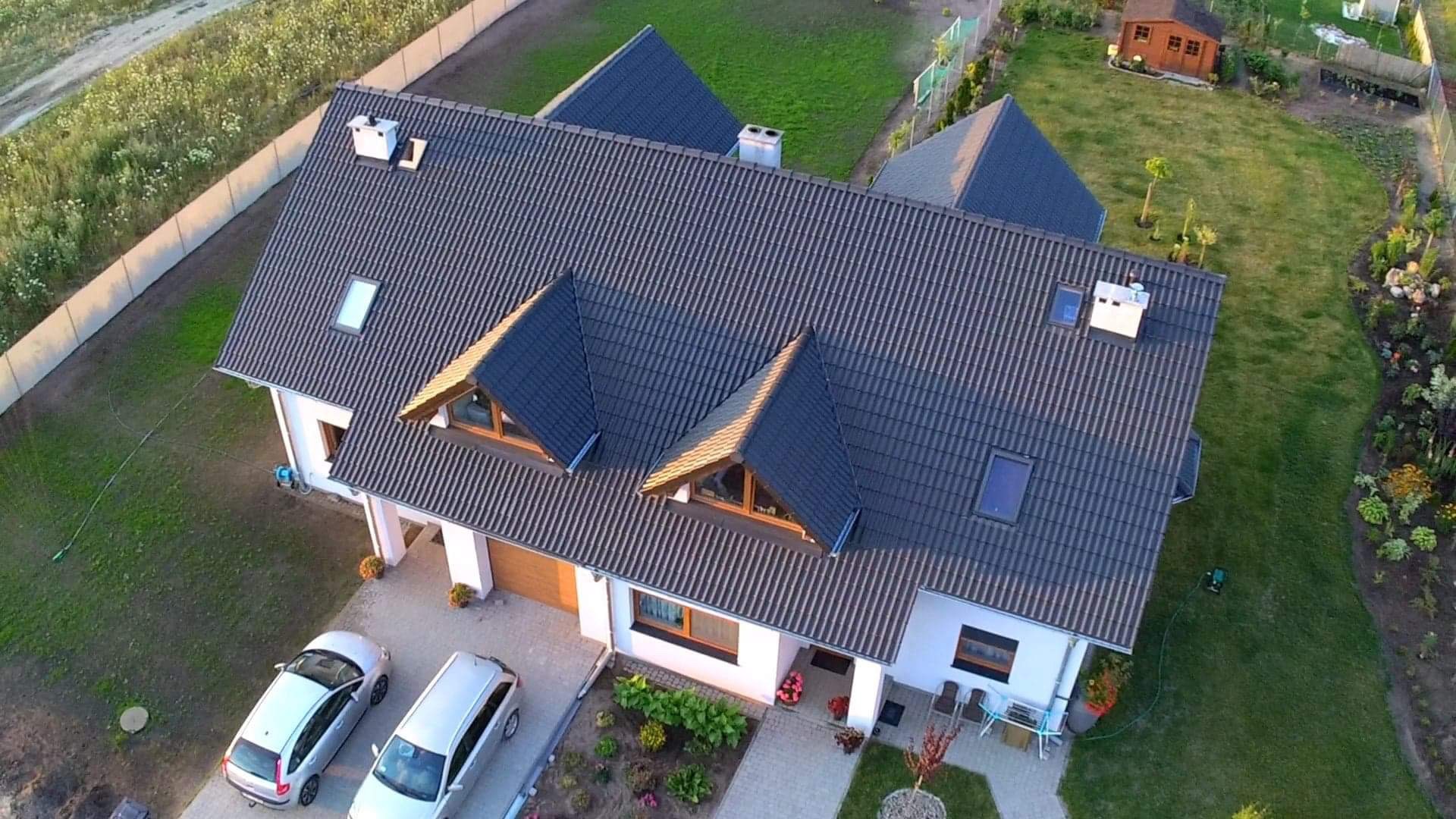 Widok z lotu ptaka na dom jednorodzinny z ciemną dachówką, lukarnami i białymi ścianami, otoczony zielonym ogrodem z dwoma samochodami zaparkowanymi na podjeździe.